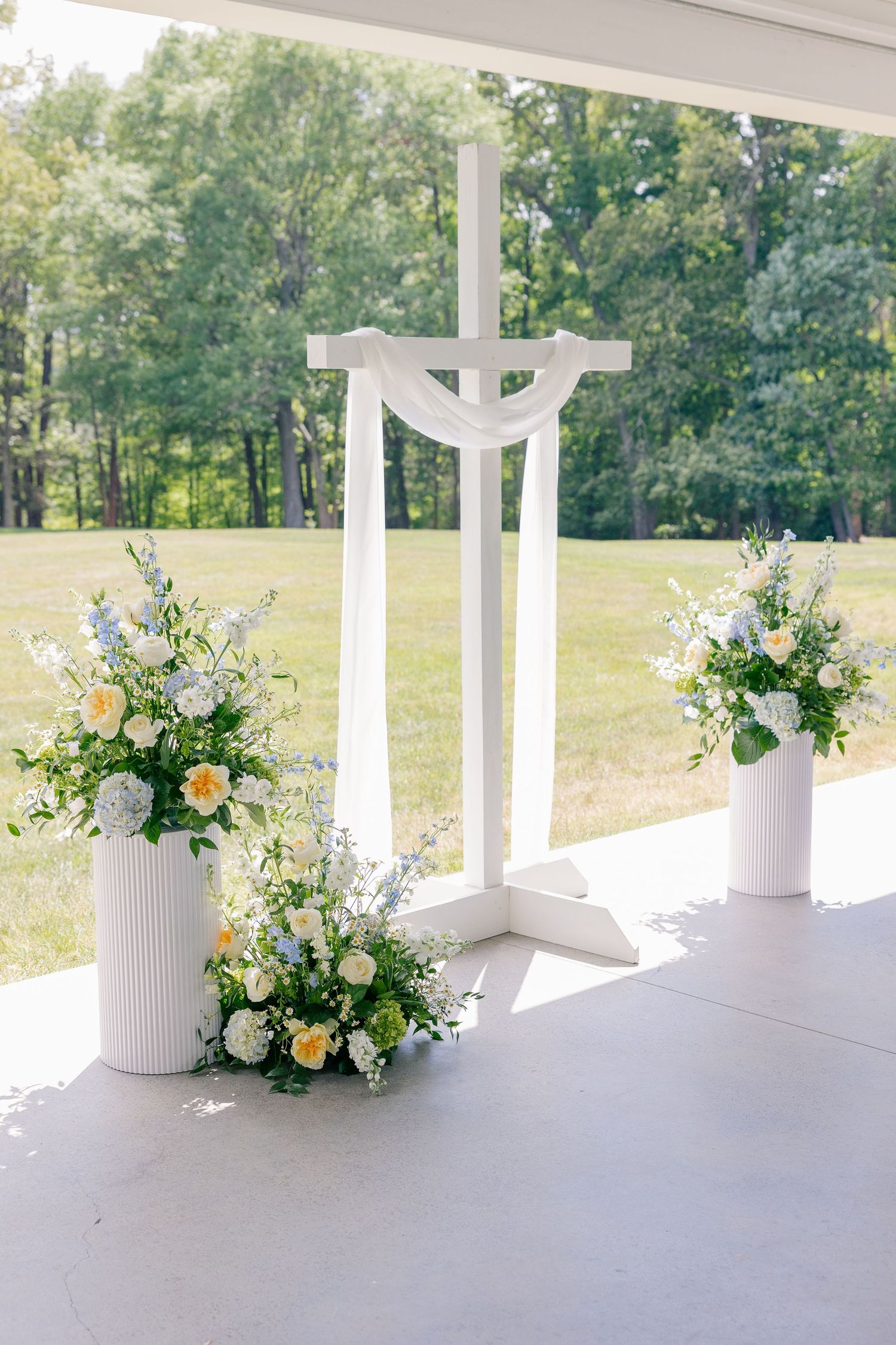 White wooden cross altar with yellow and blue floral arrangements at an outdoor wedding ceremony.