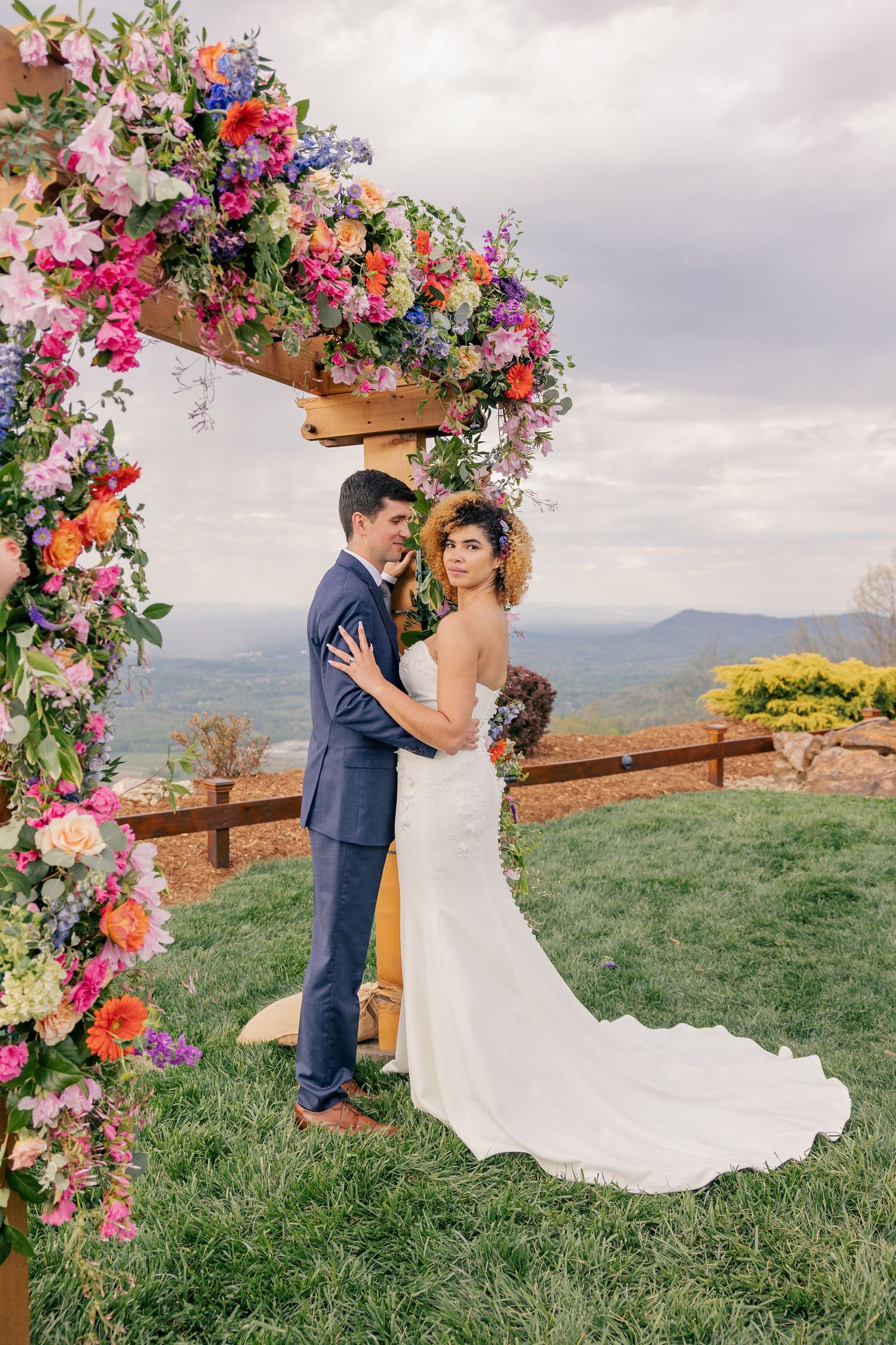 Bride and groom embrace under colorful floral arch with mountain valley views at outdoor wedding.