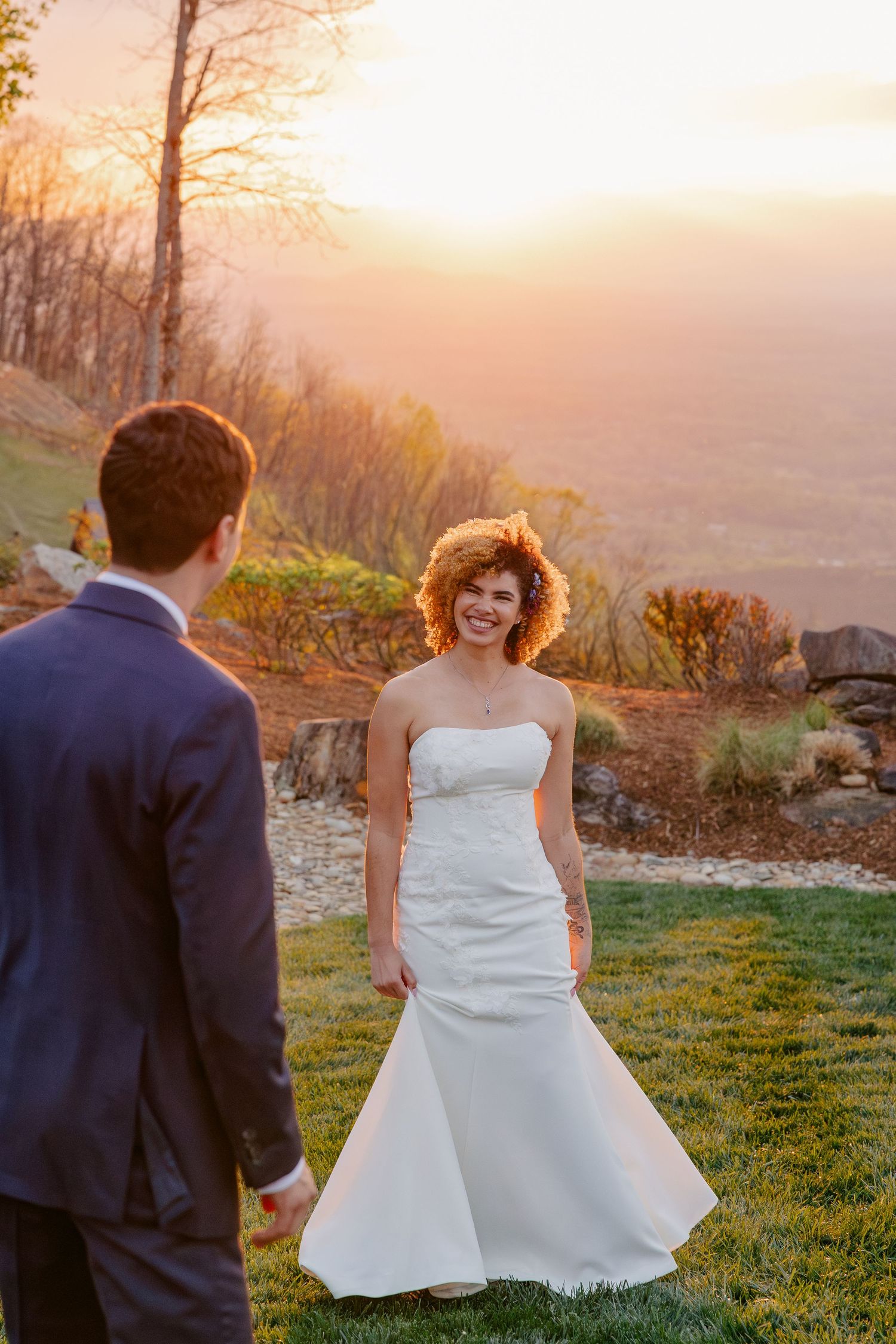 Smiling bride in white gown walks toward groom at sunset with stunning mountain valley views behind her.