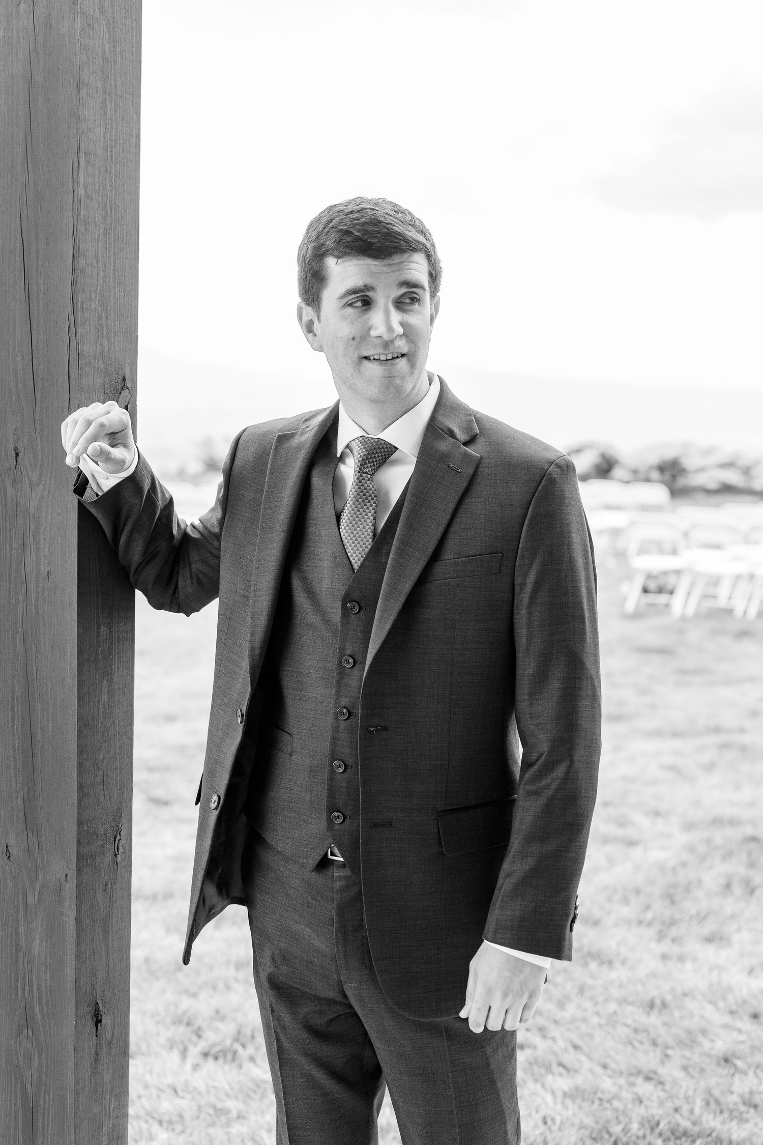 Groom in three-piece suit leans against a wooden post in a black and white outdoor wedding portrait.