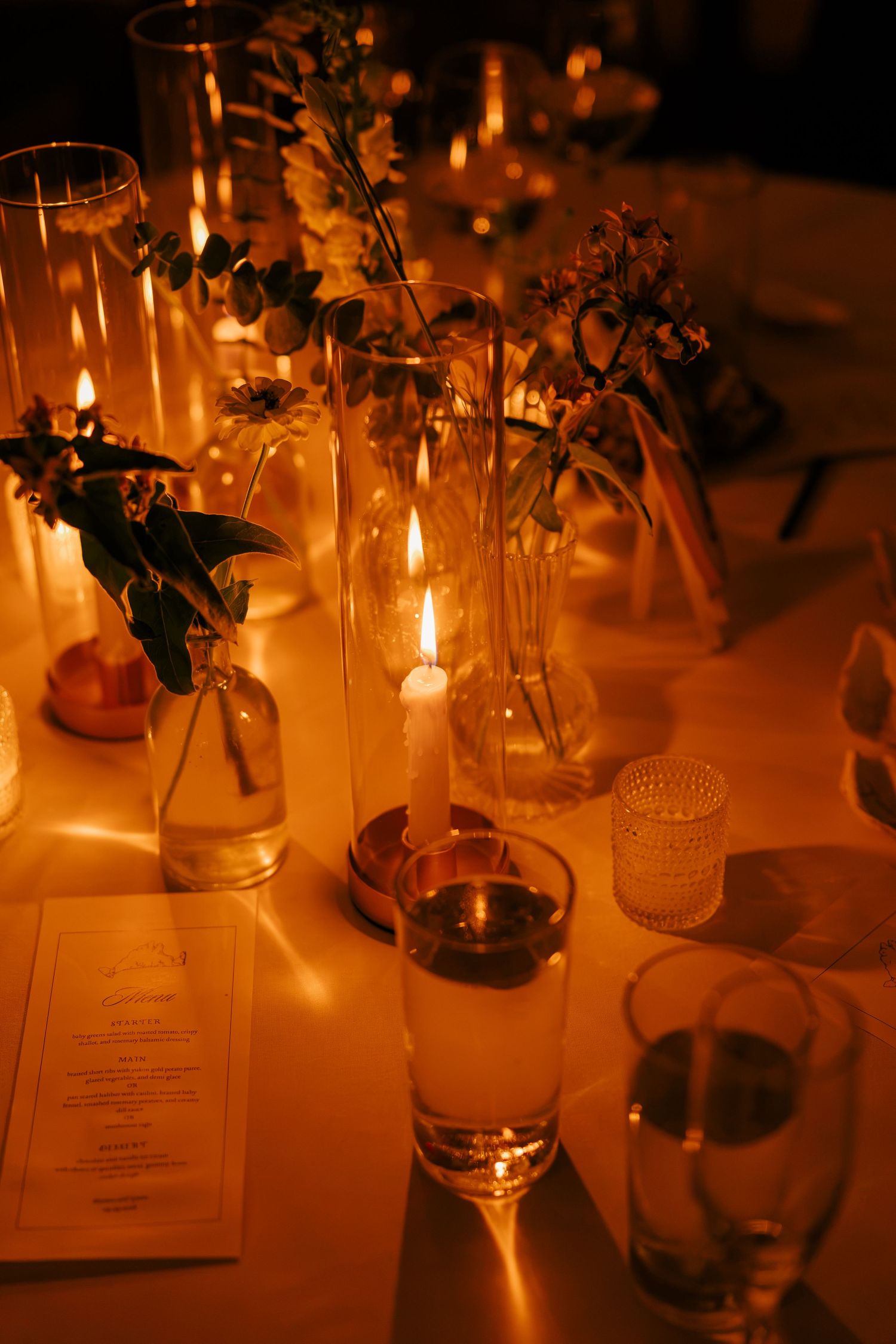 Elegant candlelit dinner table with glass vases, fresh flowers, candles, and a menu card in warm golden light.