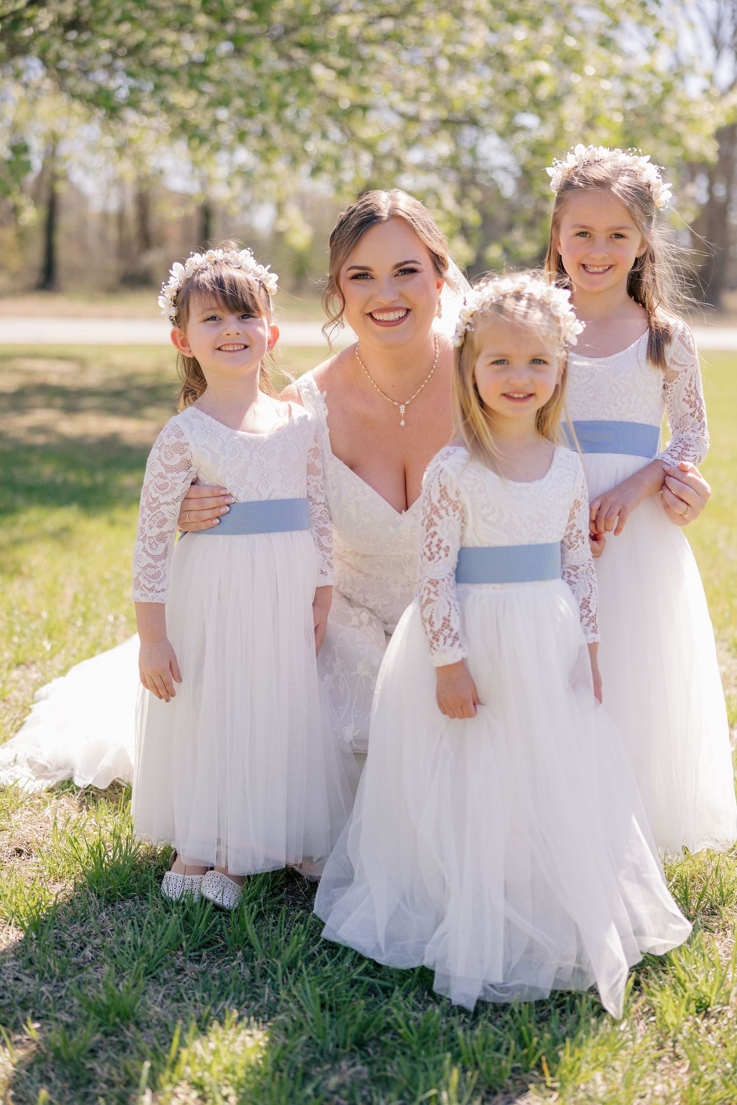 Flower girls in white lace dresses pose together on green grass in springtime.