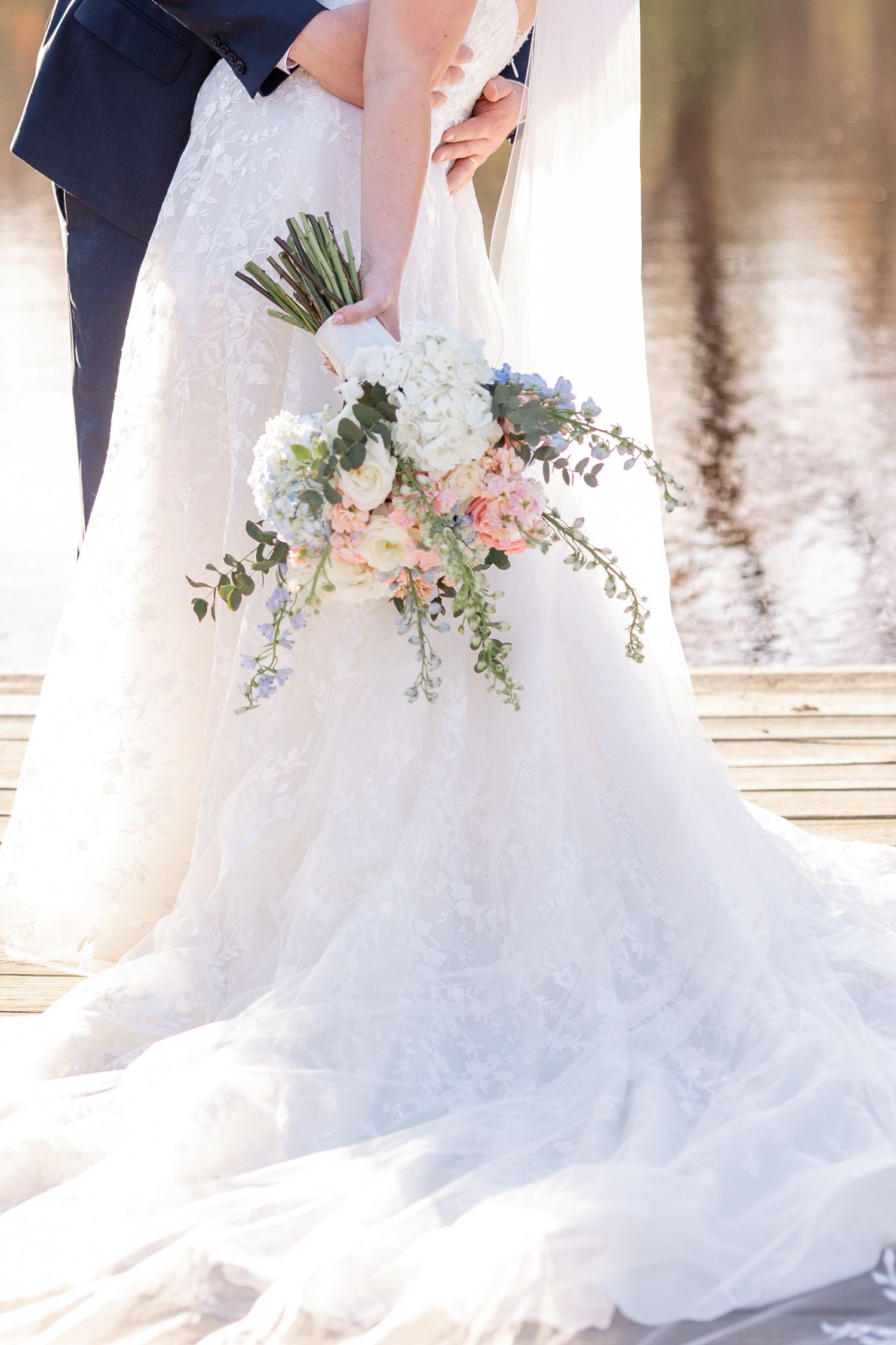 Close-up of an elegant white wedding dress train and bouquet with pink and blue flowers by the water.