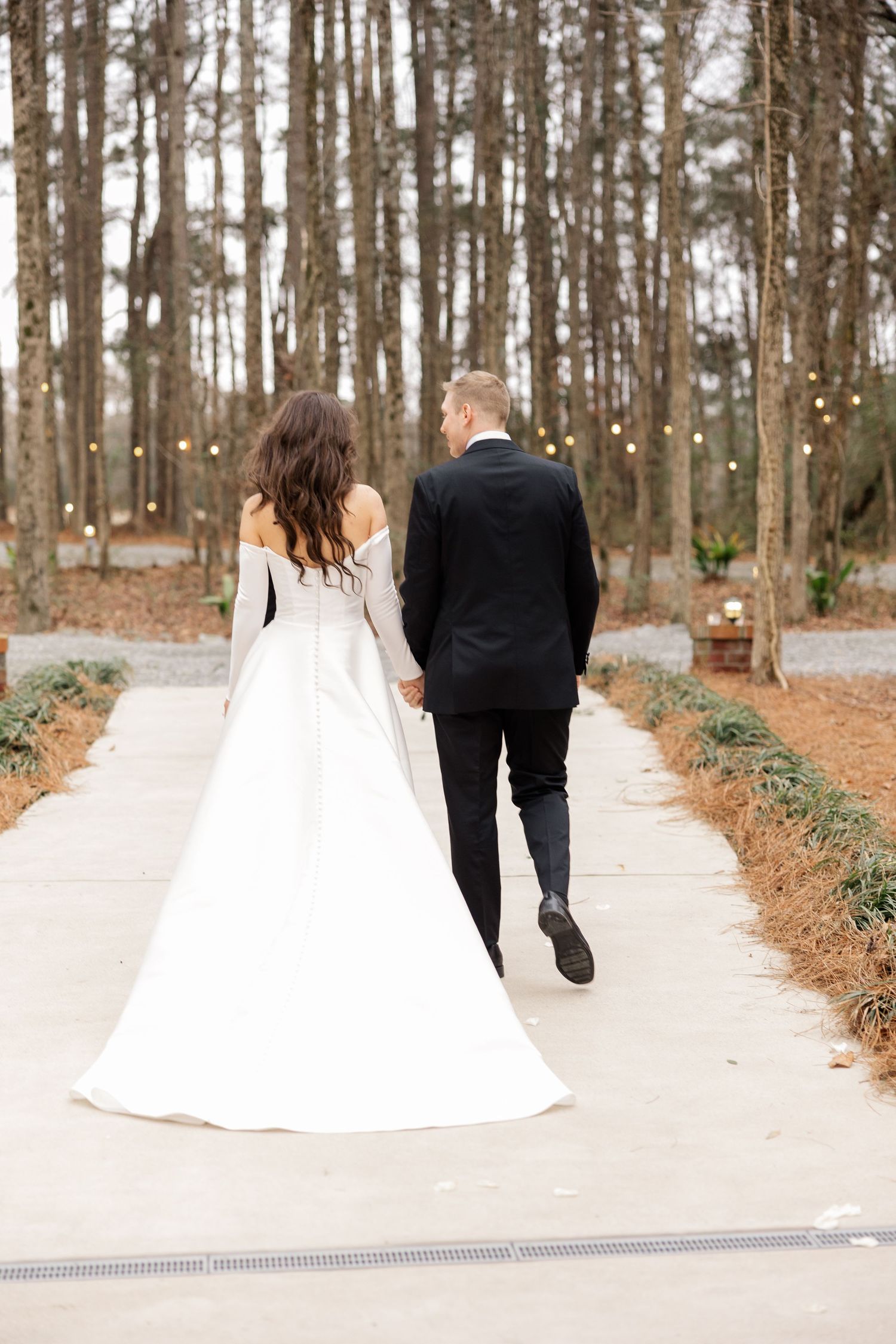 A couple walks down a winding path surrounded by tall pine trees at their winter wedding.