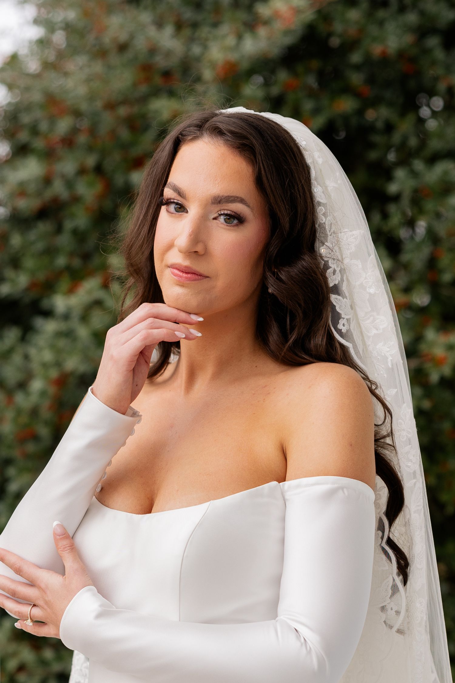 Elegant bridal portrait with white off-shoulder gown and flowing veil against a natural green backdrop.