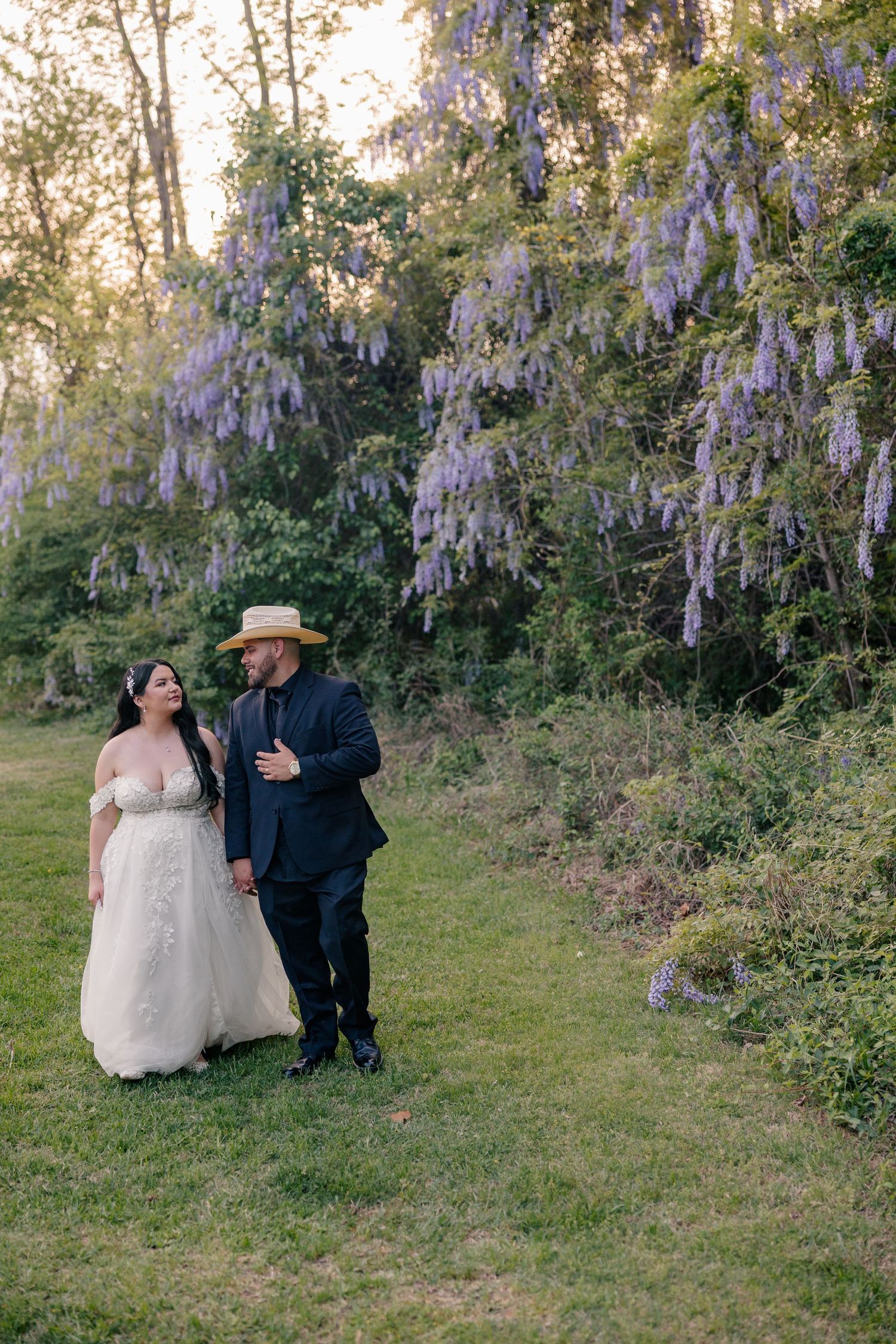 Wedding couple shares tender moments under blooming wisteria in a garden setting.