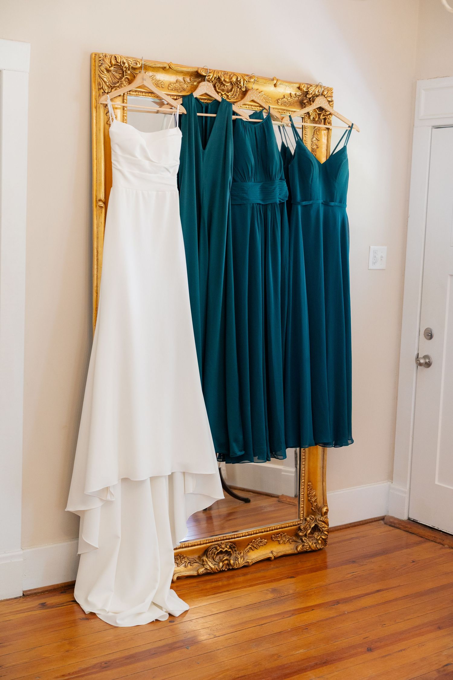 White wedding dress and teal bridesmaid dresses hang elegantly against an ornate gold-framed mirror.