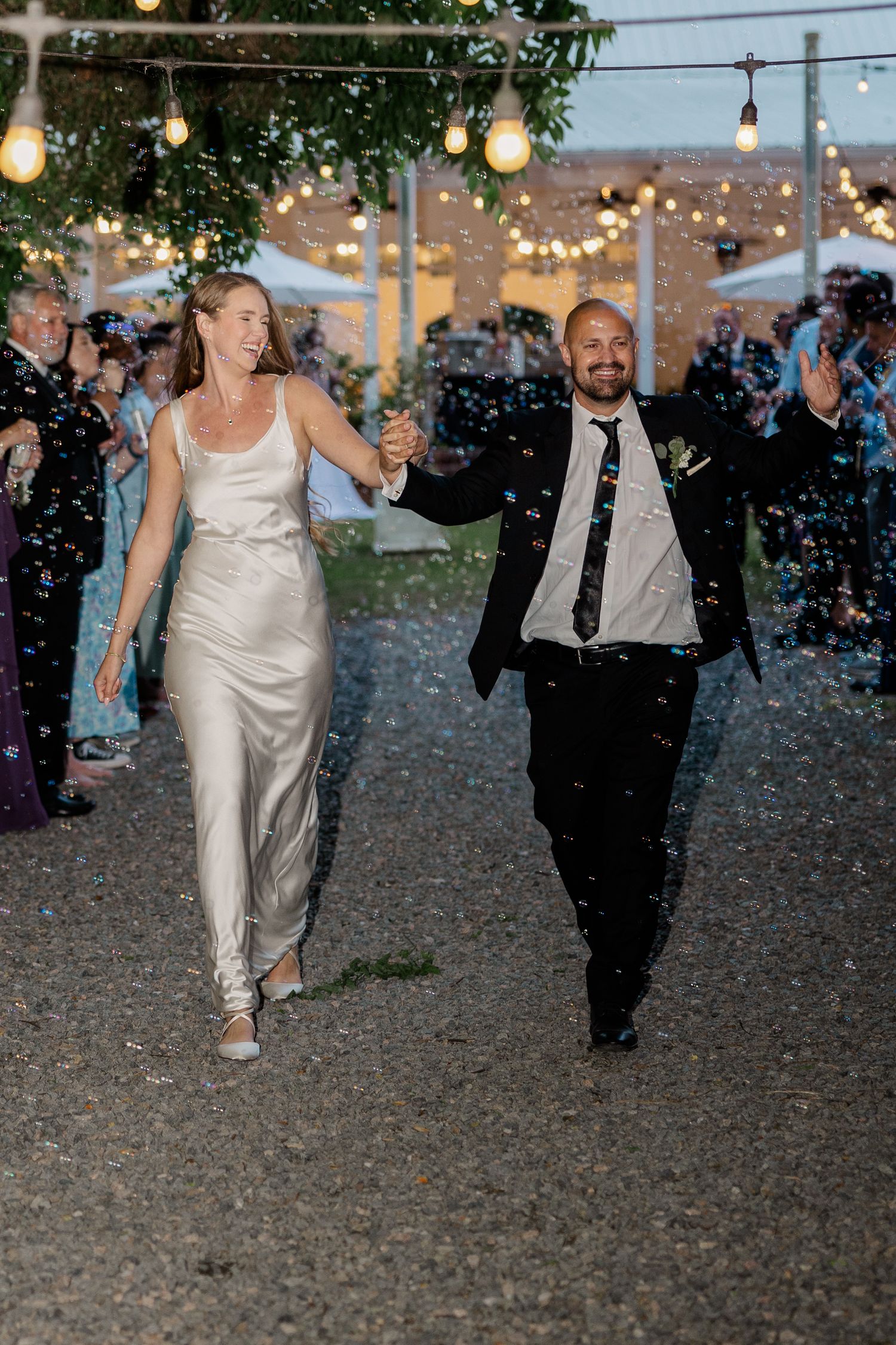 Newlyweds exit their reception under twinkling string lights while guests celebrate.