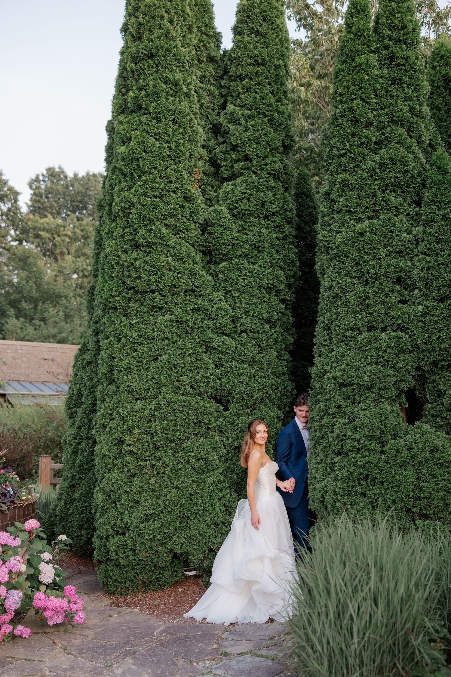A wedding couple poses for portraits in a garden setting with tall evergreen trees and pink flowers.