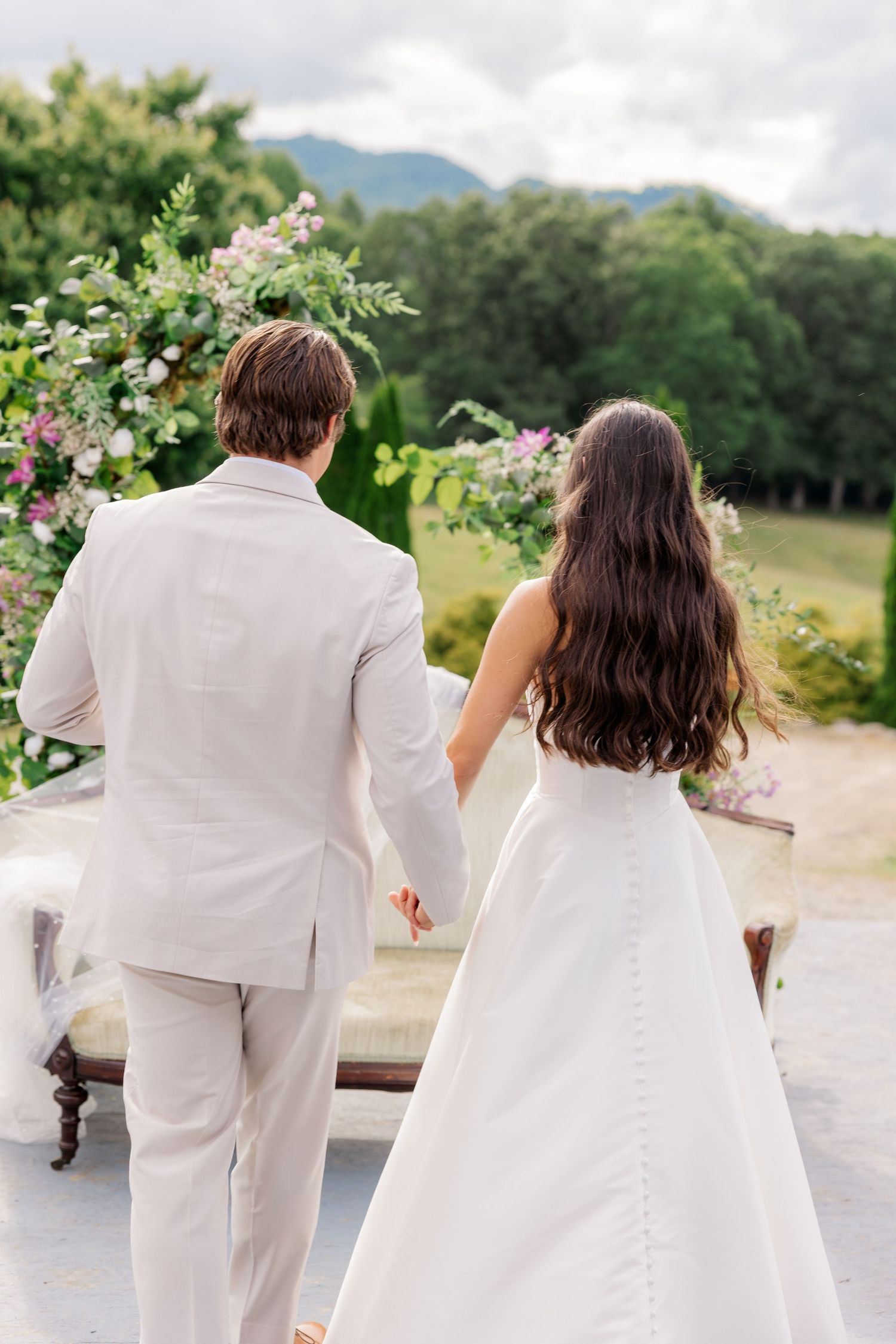 A couple in white attire walking together through a scenic outdoor setting with mountains and flowers in the background.