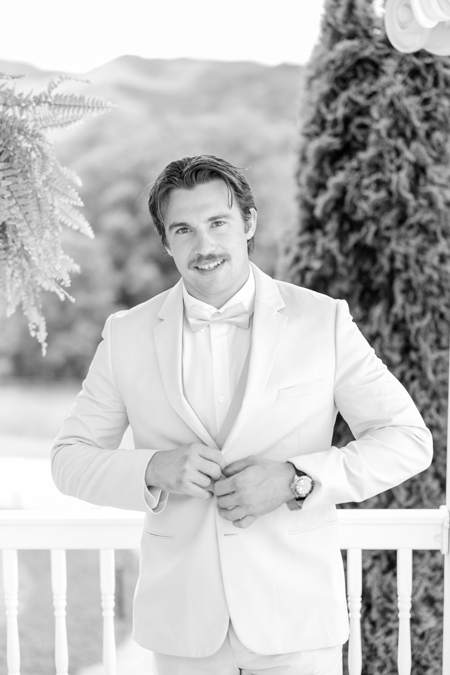 A groom in a crisp white tuxedo jacket and bow tie poses outdoors against a railing on his wedding day.