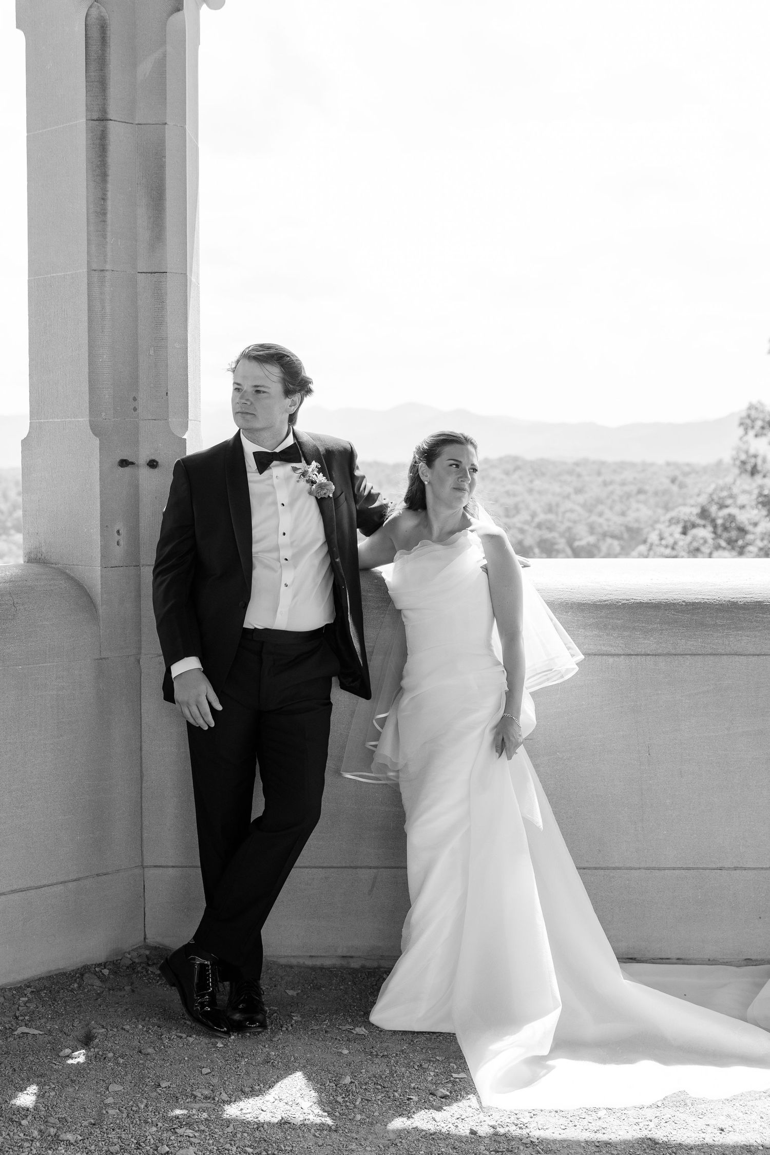 Bride and groom pose in black and white portrait on stone balcony with mountain views behind them.