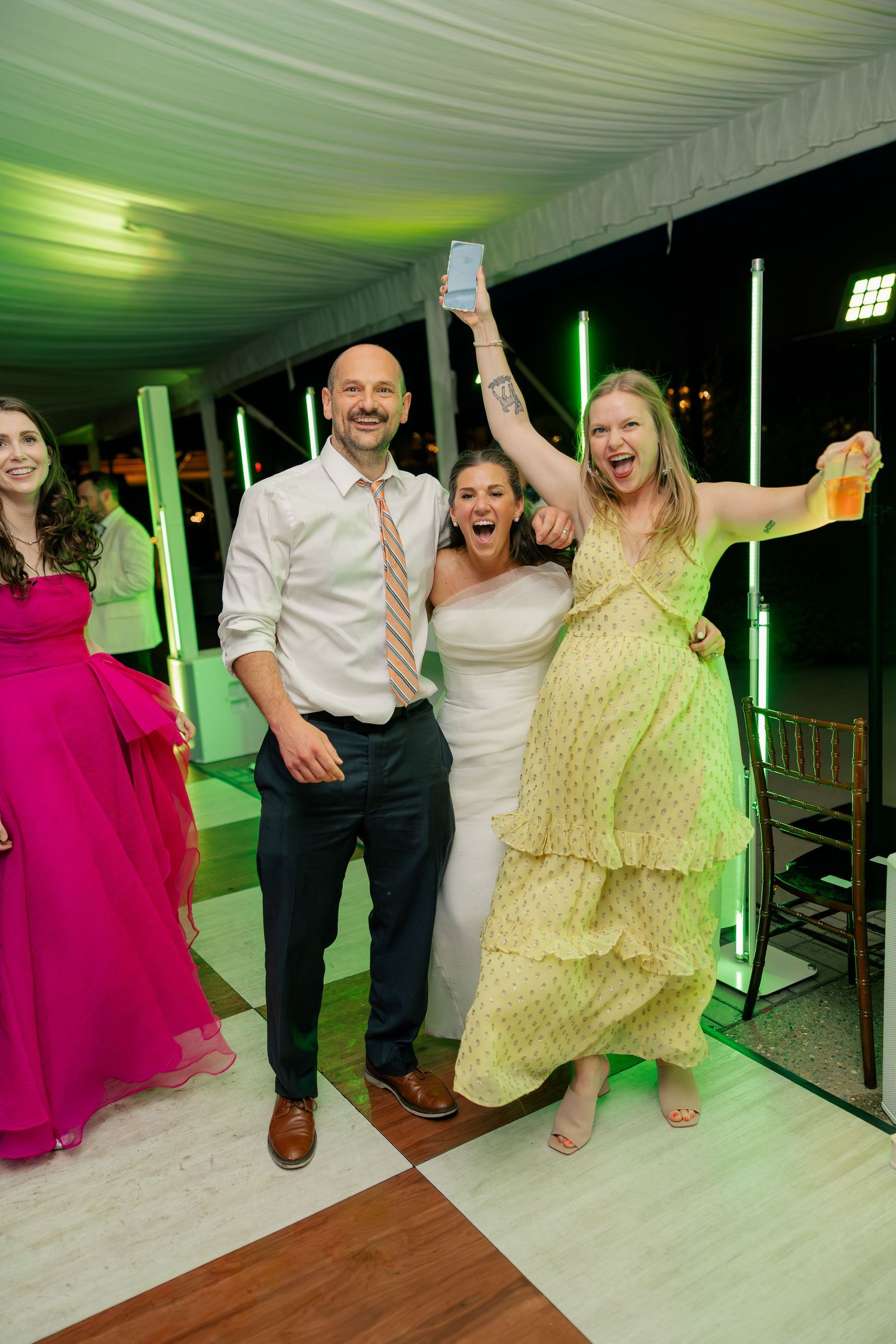 Joyful bride and guests celebrate on dance floor under green lights at wedding reception.