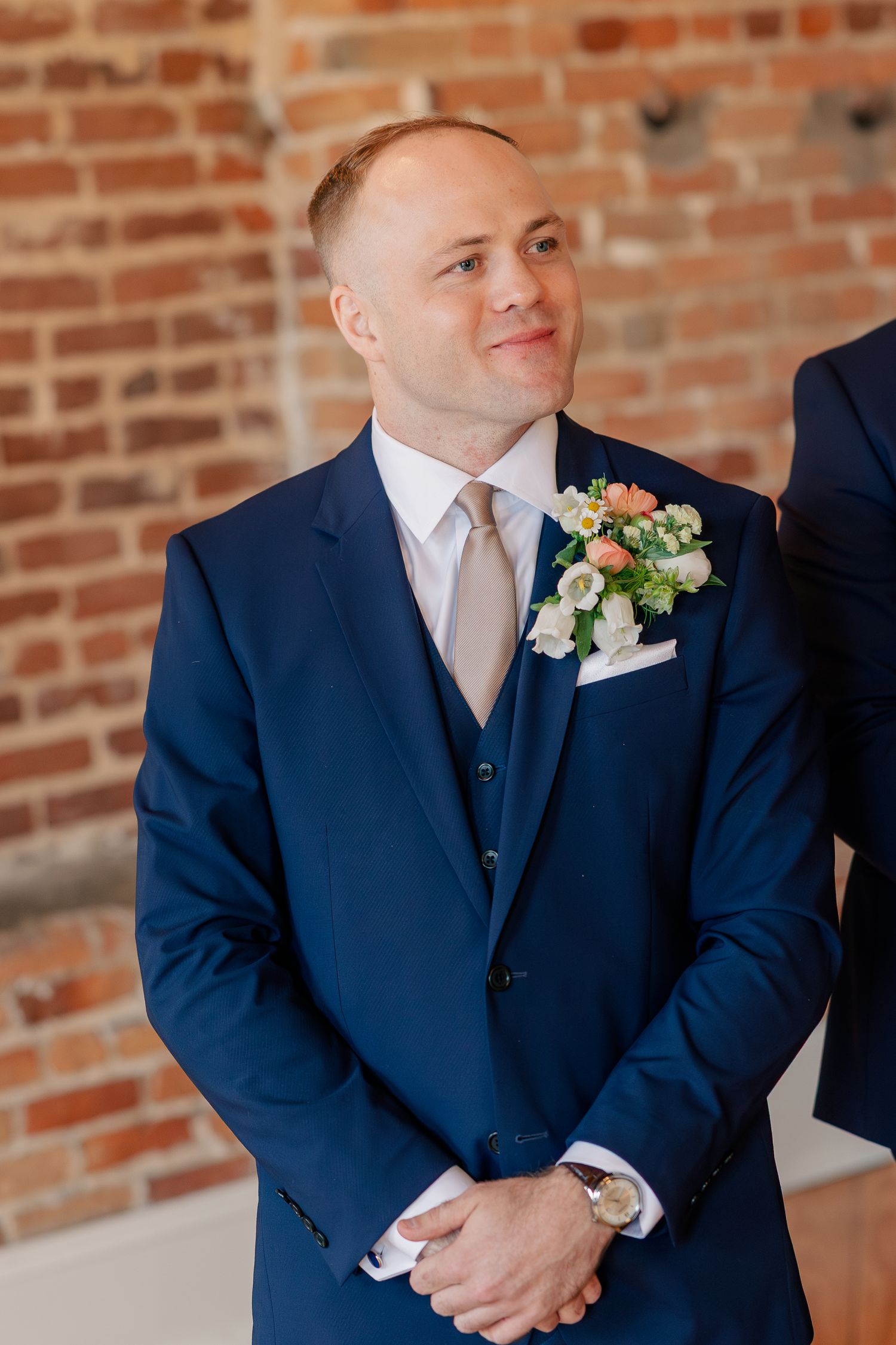 Dapper groom in navy three-piece suit with floral boutonniere smiles during wedding ceremony.