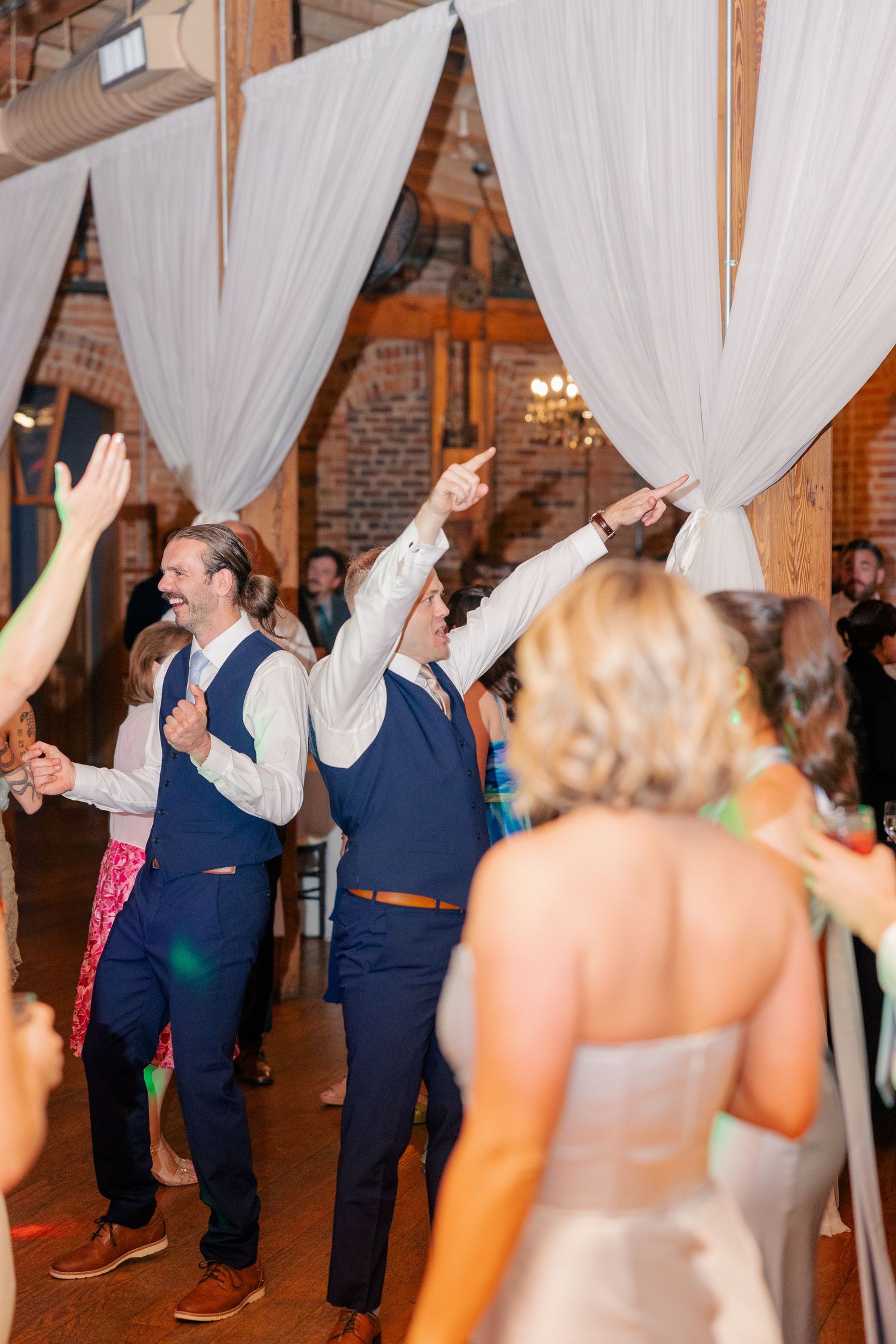 Two groomsmen in navy vests celebrate and point upward on the dance floor during a lively wedding reception.
