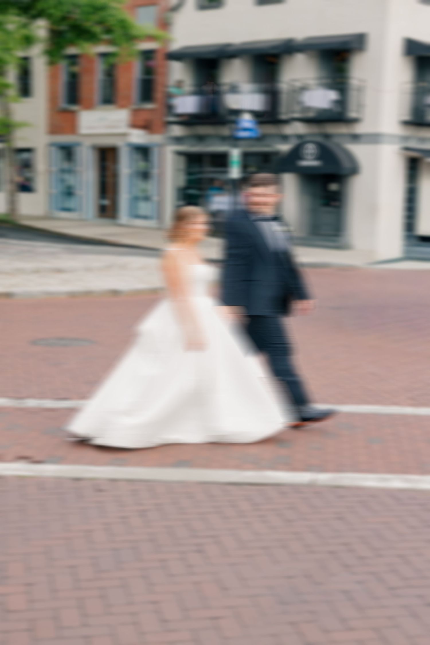 Blurred motion shot of bride in white gown and groom crossing brick street.