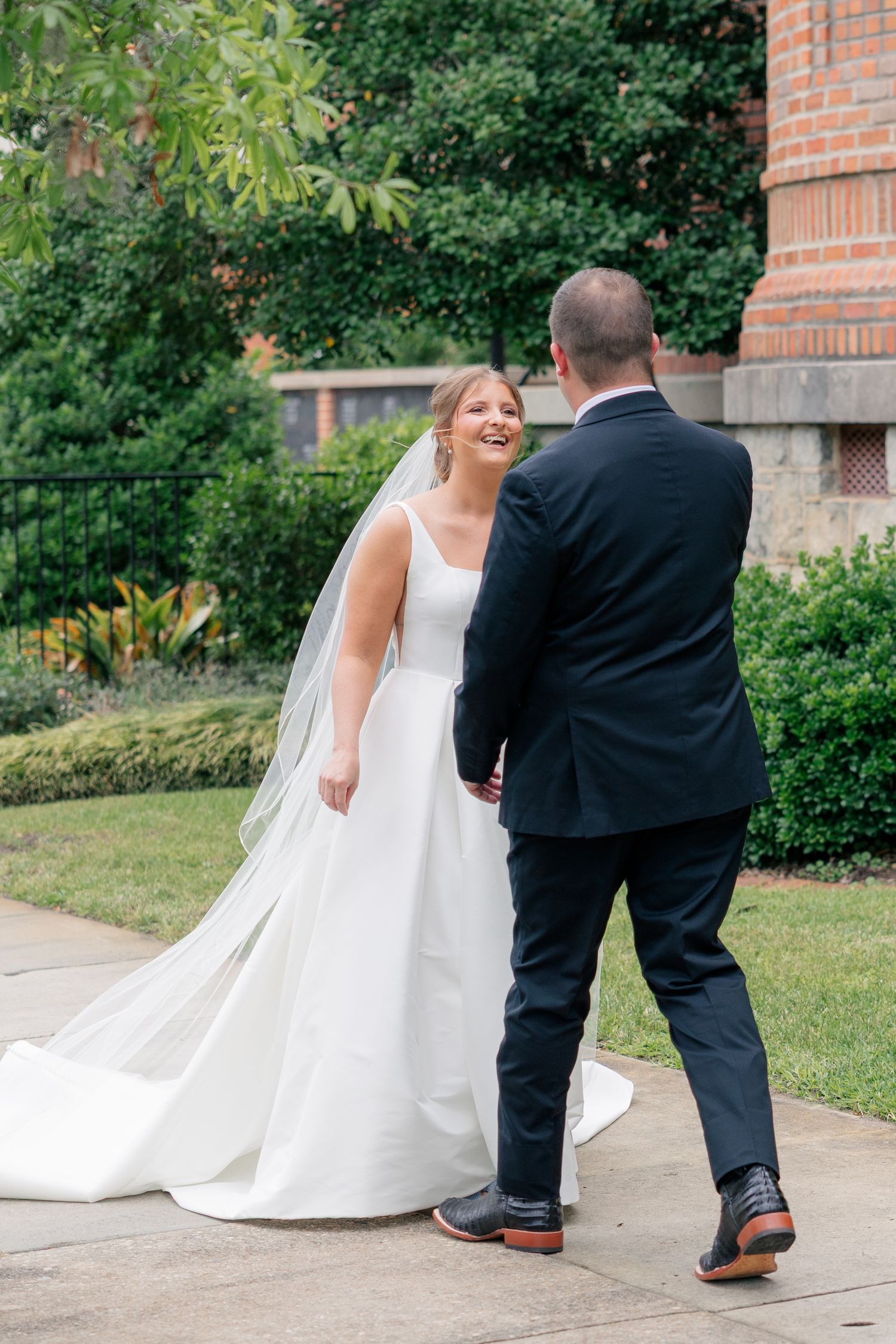 Bride in a white gown and veil laughs with groom in navy suit during first look outside brick building.
