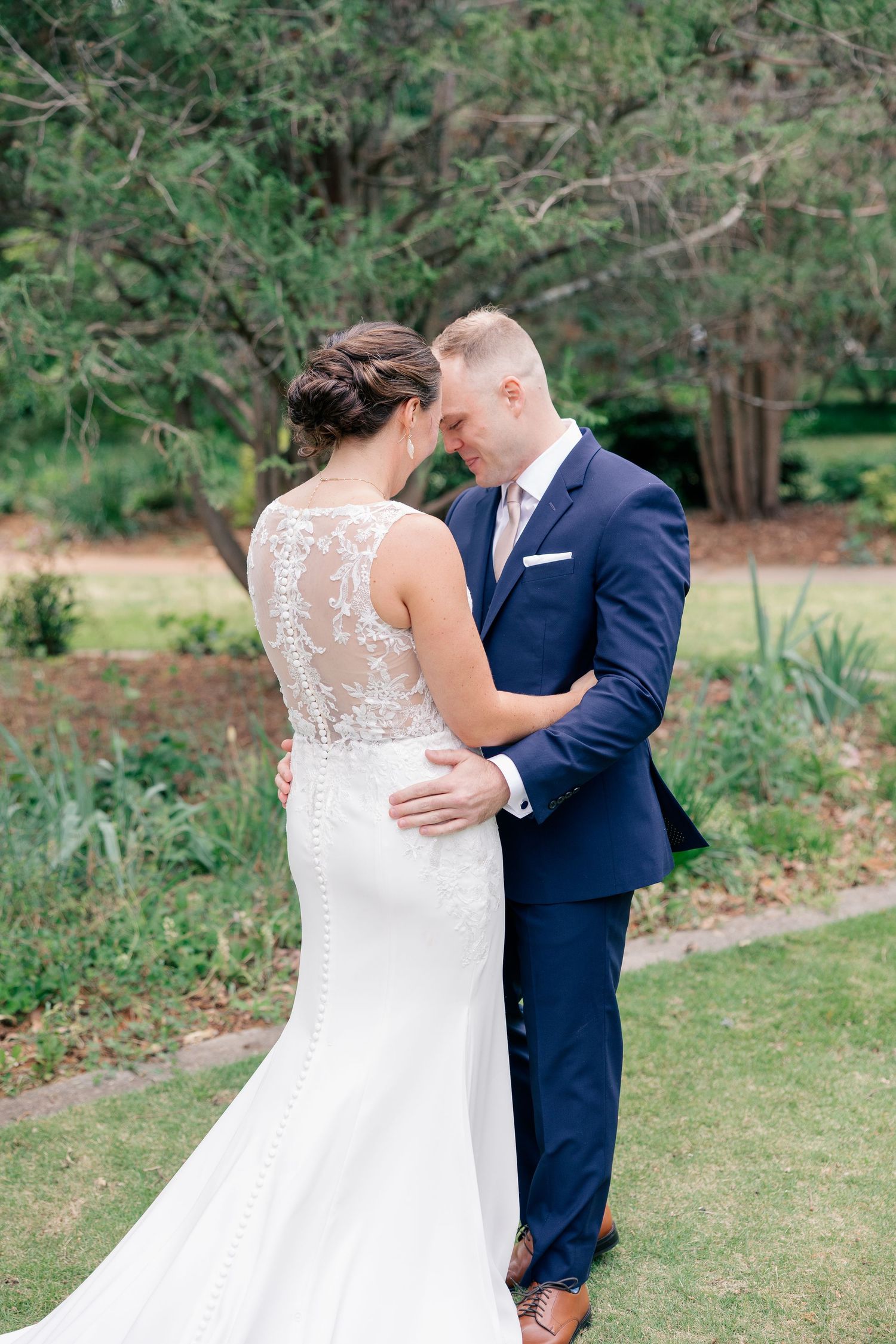 Bride and groom share tender moment in lush garden, showcasing intricate lace back wedding gown.
