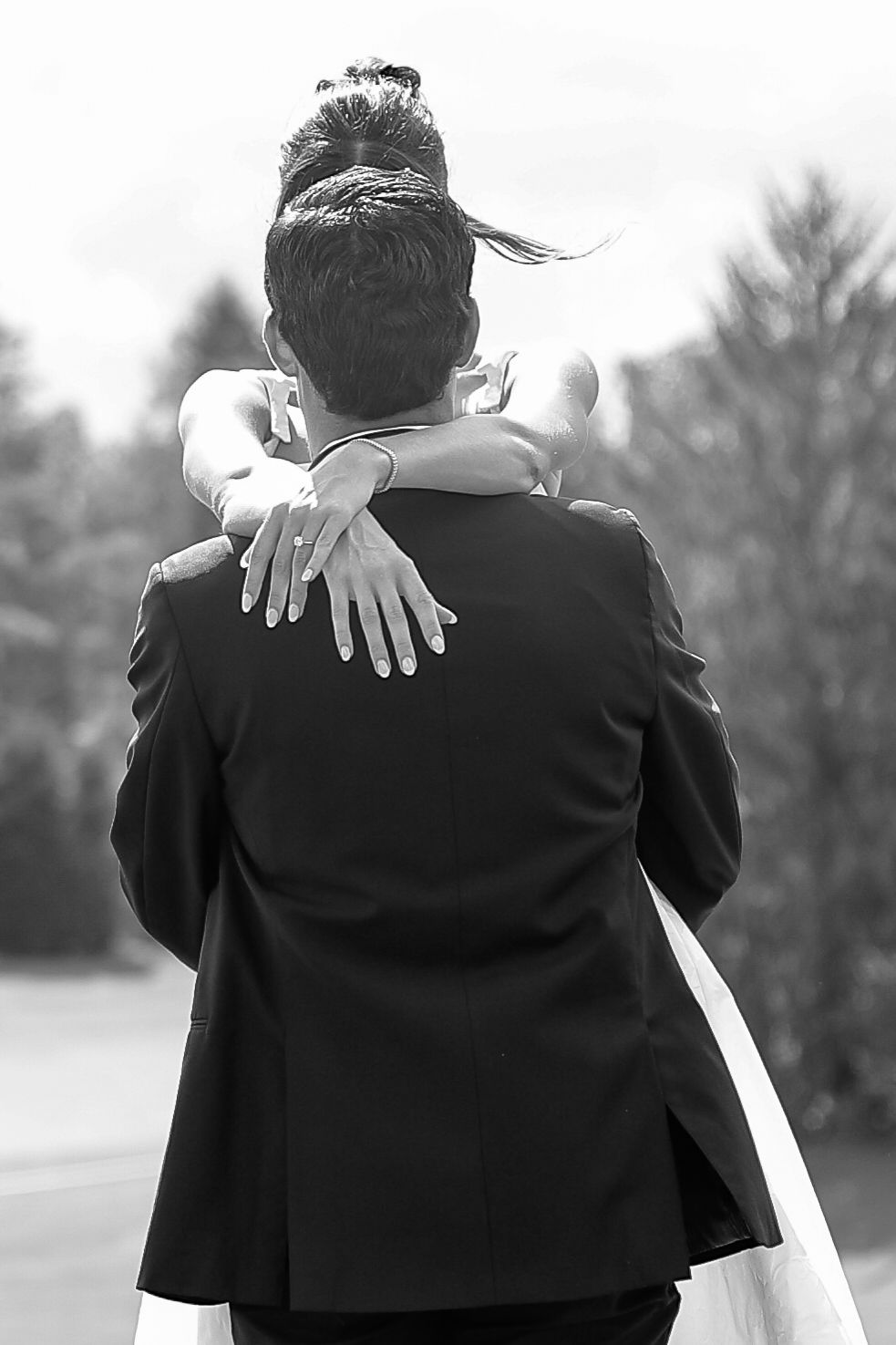 Black and white photo of bride embracing groom from behind, showing wedding ring on her hand.