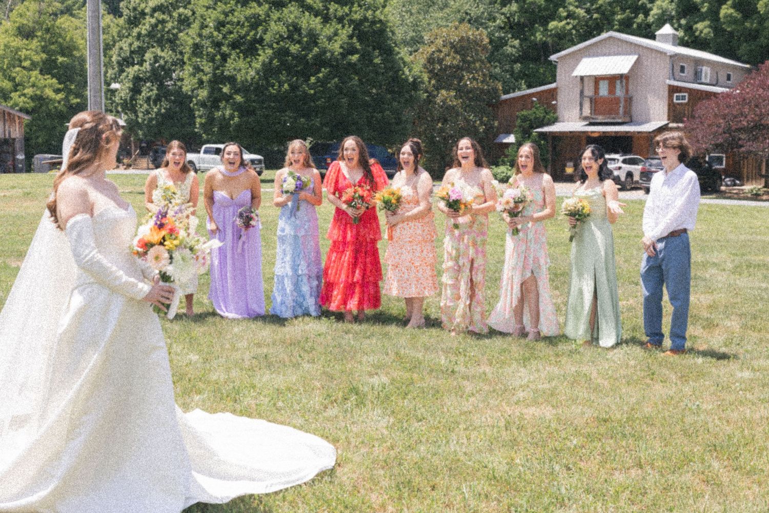 Bride in white gown faces bridesmaids in colorful mismatched dresses during outdoor wedding party reveal.