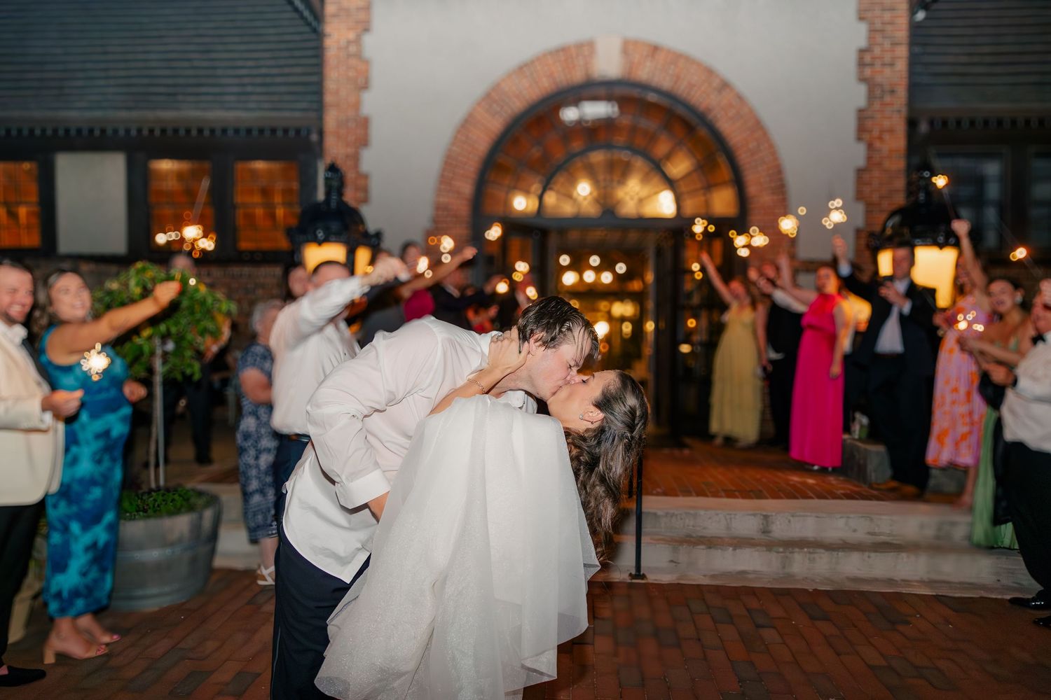 Bride and groom share romantic dip kiss during sparkler sendoff at elegant evening wedding reception.