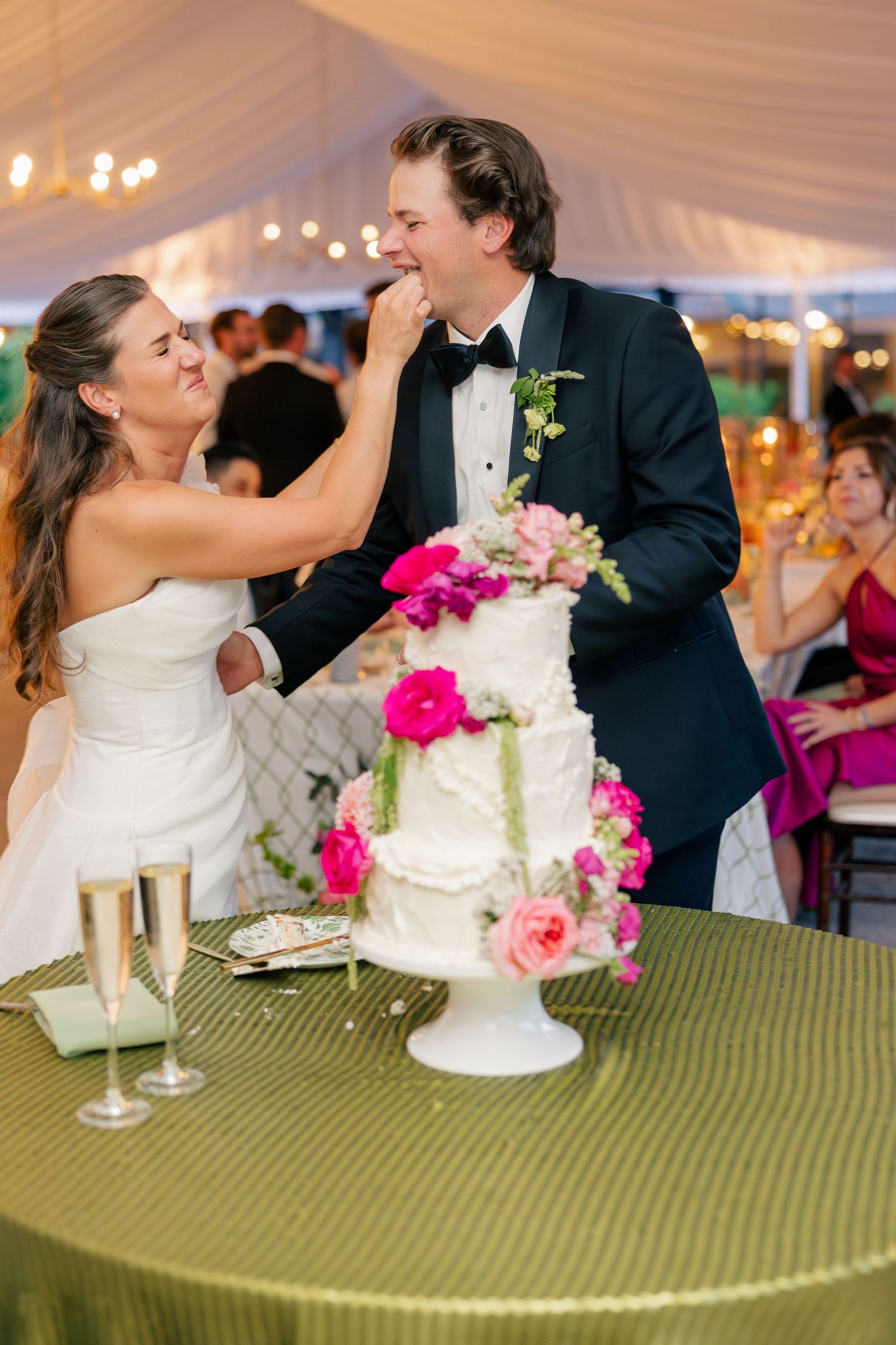 Bride feeds groom wedding cake at reception with white floral cake and champagne glasses nearby.