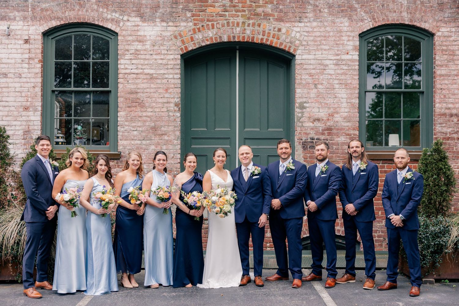 Wedding party poses in front of brick building with green doors, bridesmaids in blue gowns and groomsmen in navy suits.