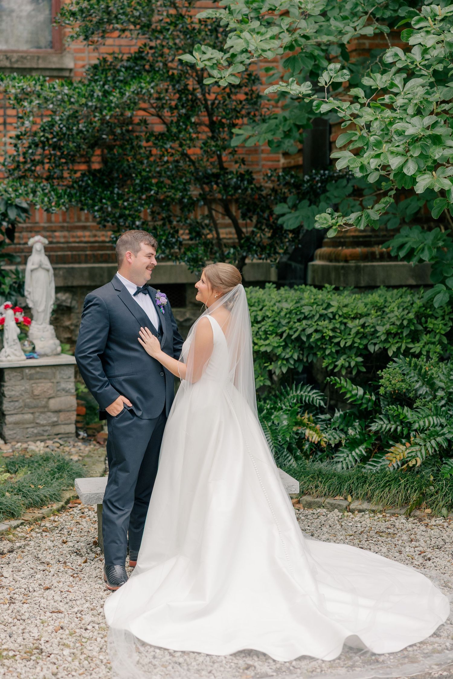 Bride and groom share romantic moment in garden courtyard, bride in flowing white gown with veil, groom in navy suit.