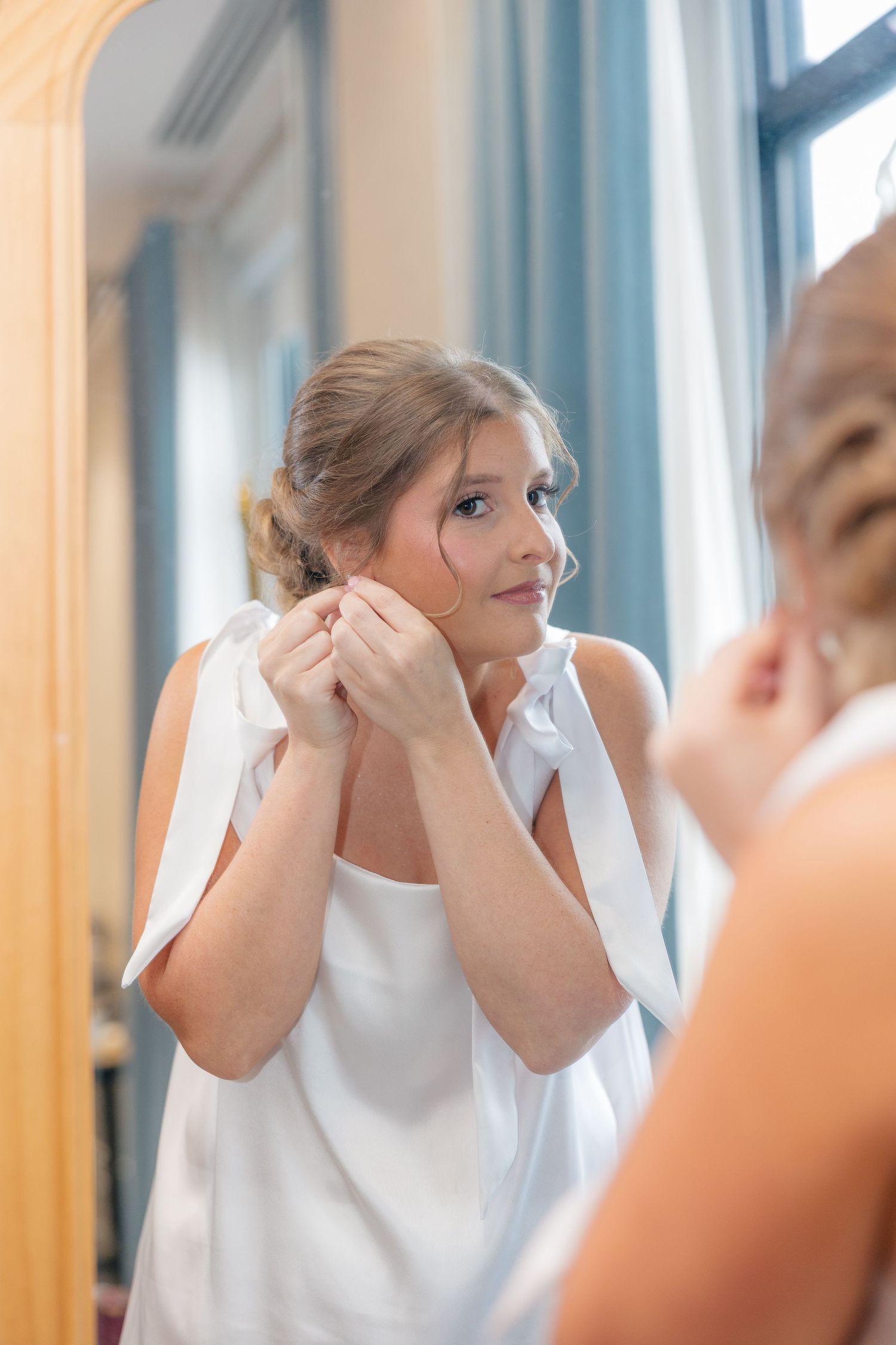Bride in white dress putting on earrings while looking in a mirror during wedding preparations.