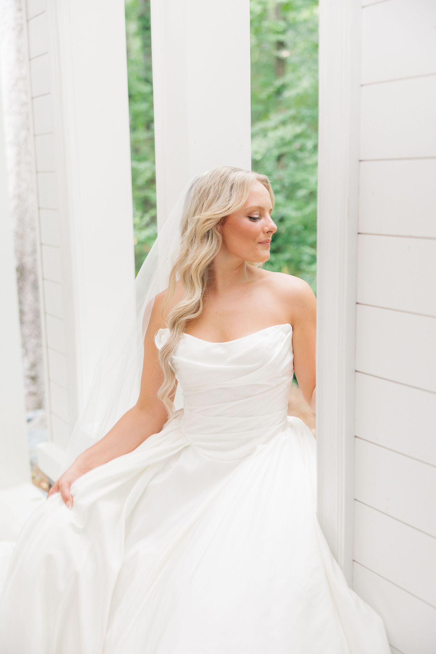 Elegant bride in strapless white ball gown sits between white columns with soft natural lighting.