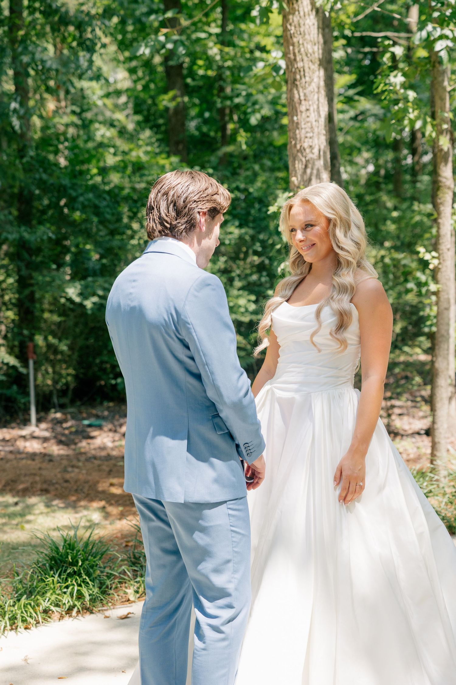 Bride in white gown smiles at groom in light blue suit during outdoor first look ceremony surrounded by trees.