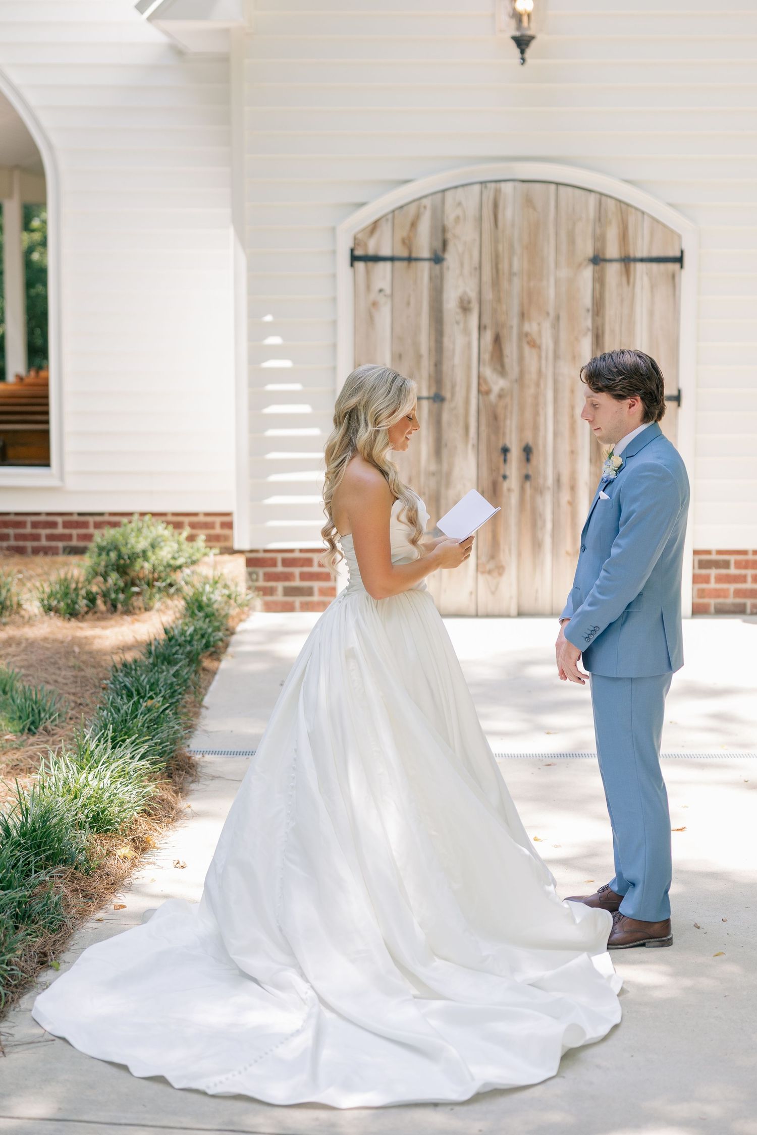 Bride reading vows to groom during intimate first look outside white chapel with wooden barn doors.