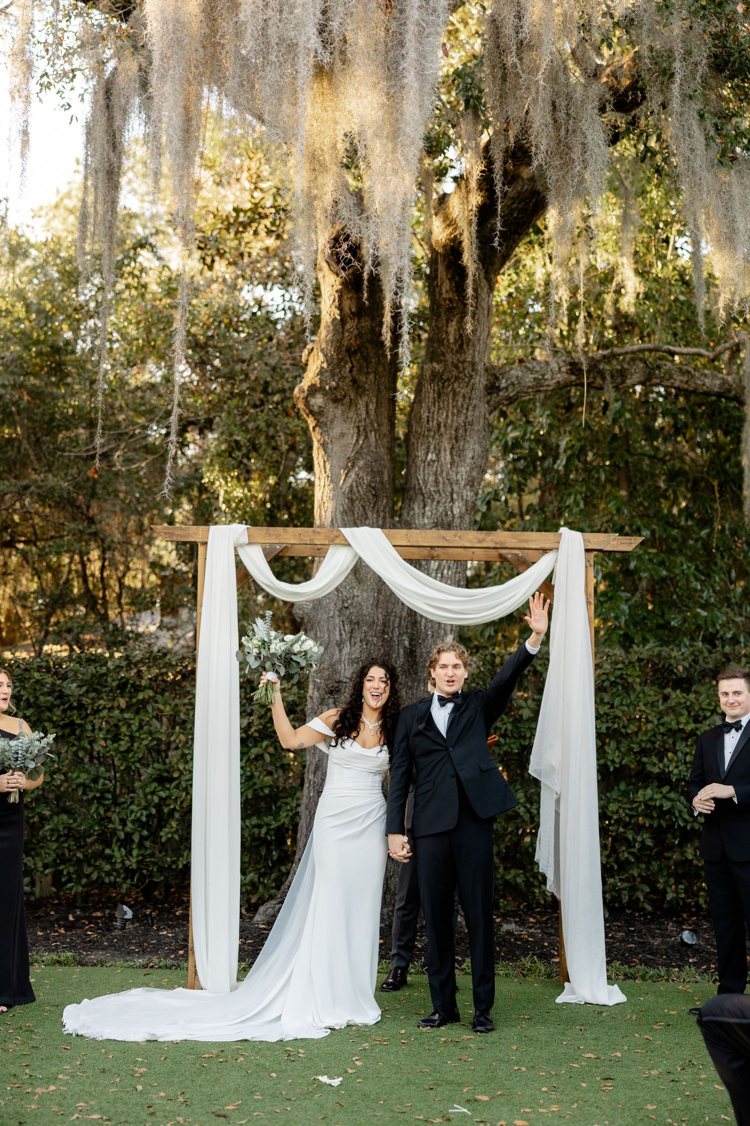 A newlywed couple celebrates under a white fabric arch decorated with Spanish moss.