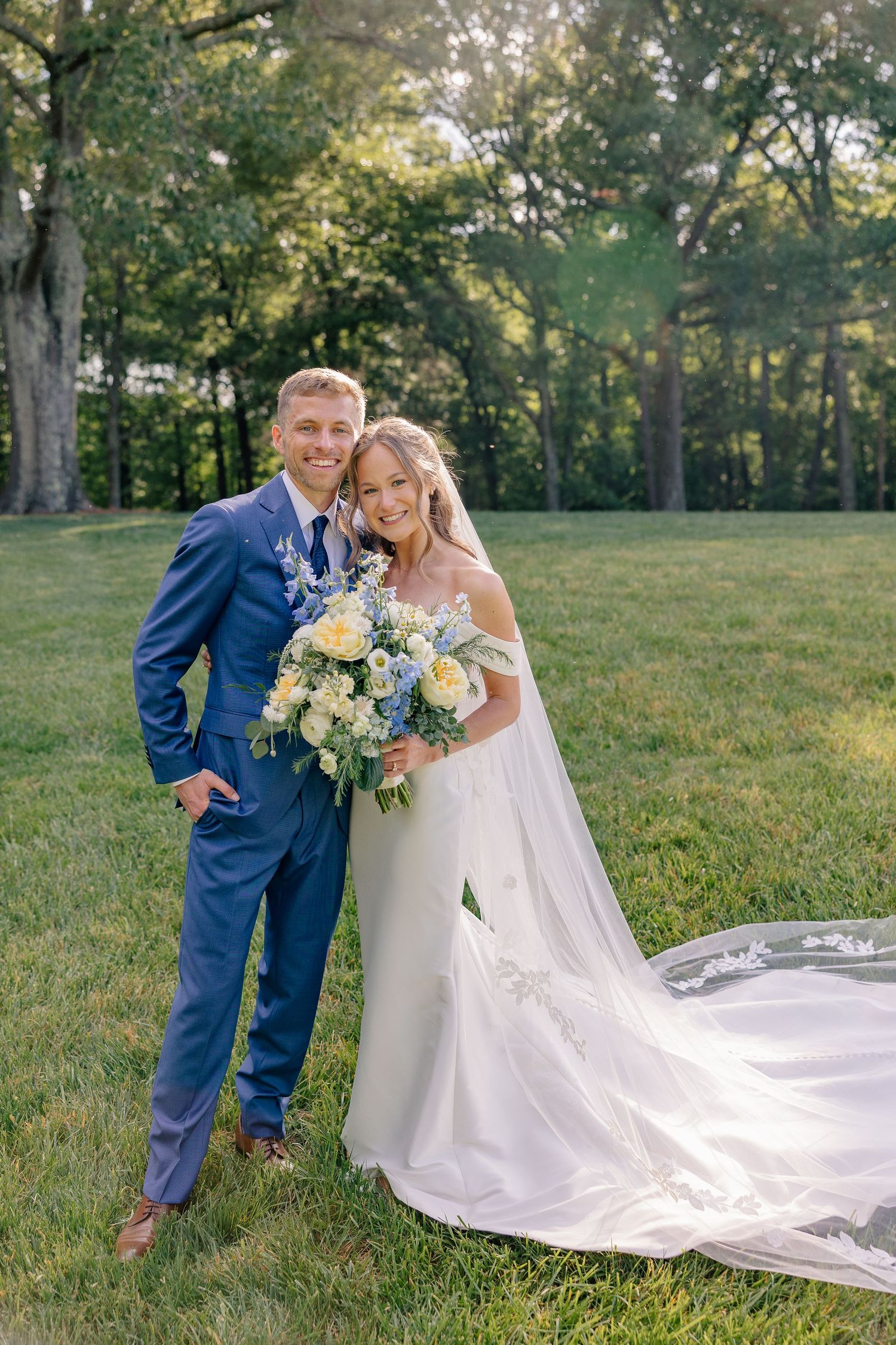 A happy bride and groom pose together outdoors, the bride holding a bouquet of blue and yellow flowers.