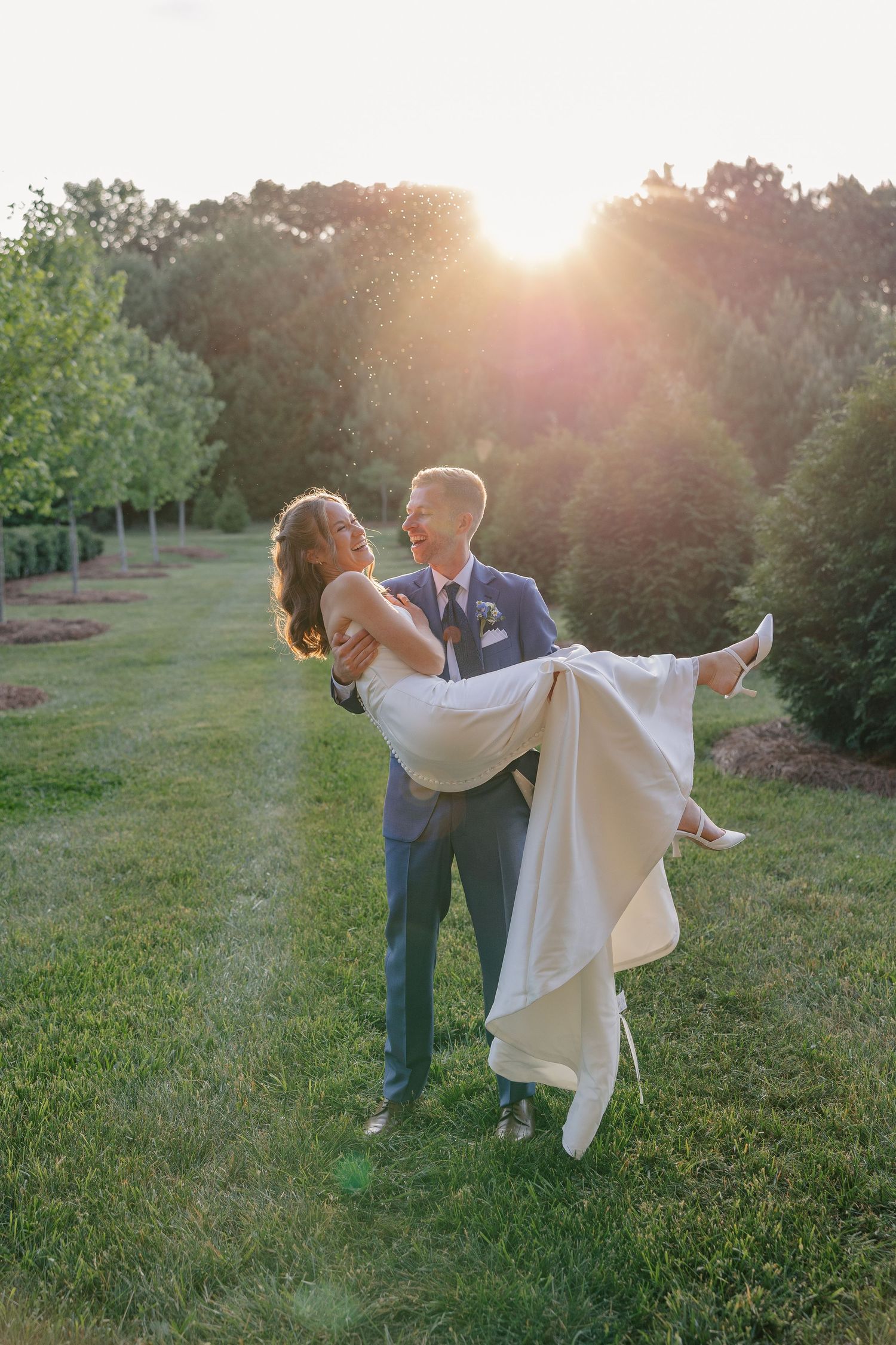 Groom lifts smiling bride in wedding dress during stunning golden hour outdoor photo.
