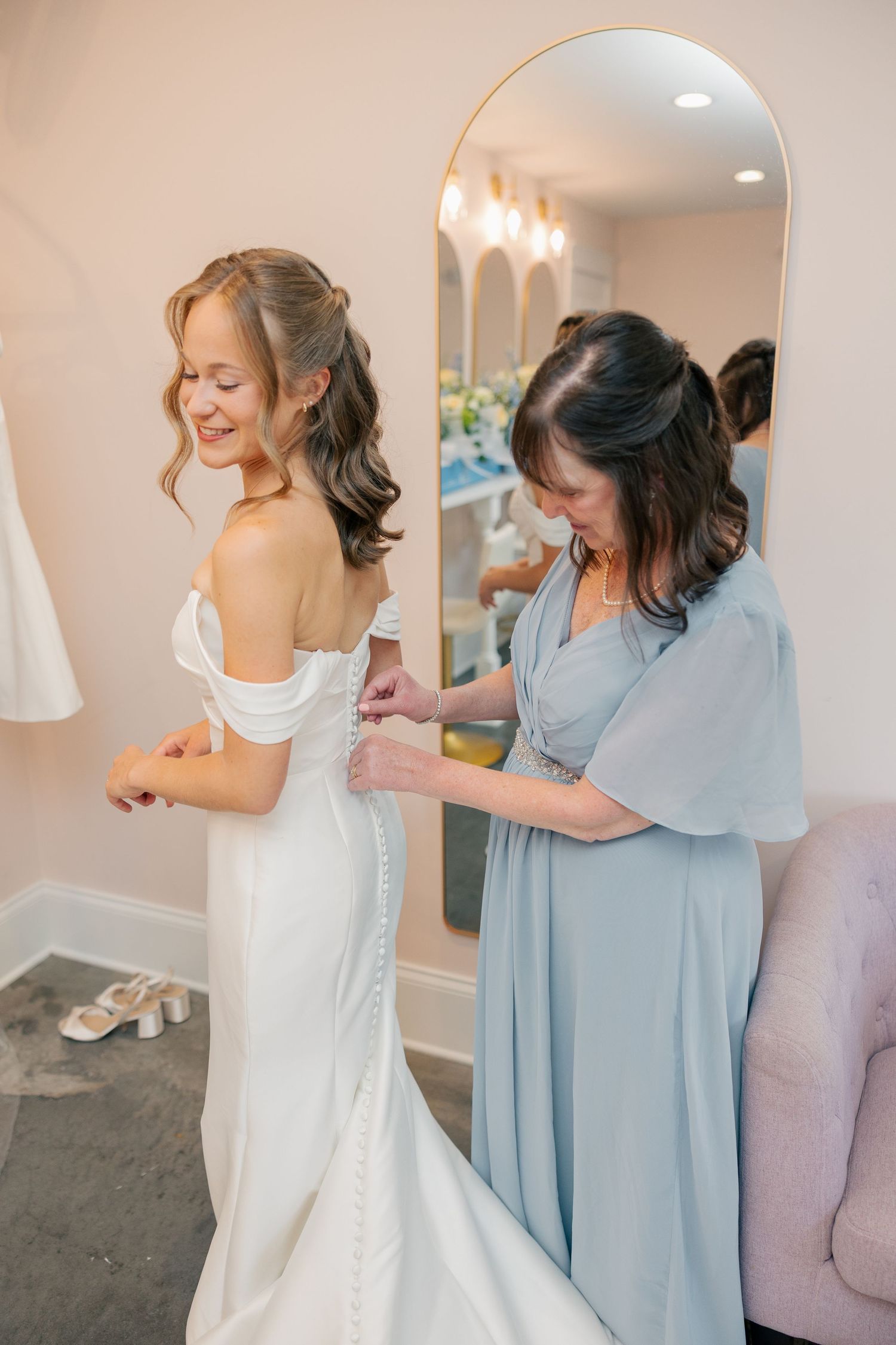 A mother helps her smiling bride button the back of her white off-shoulder wedding gown in a bridal suite.