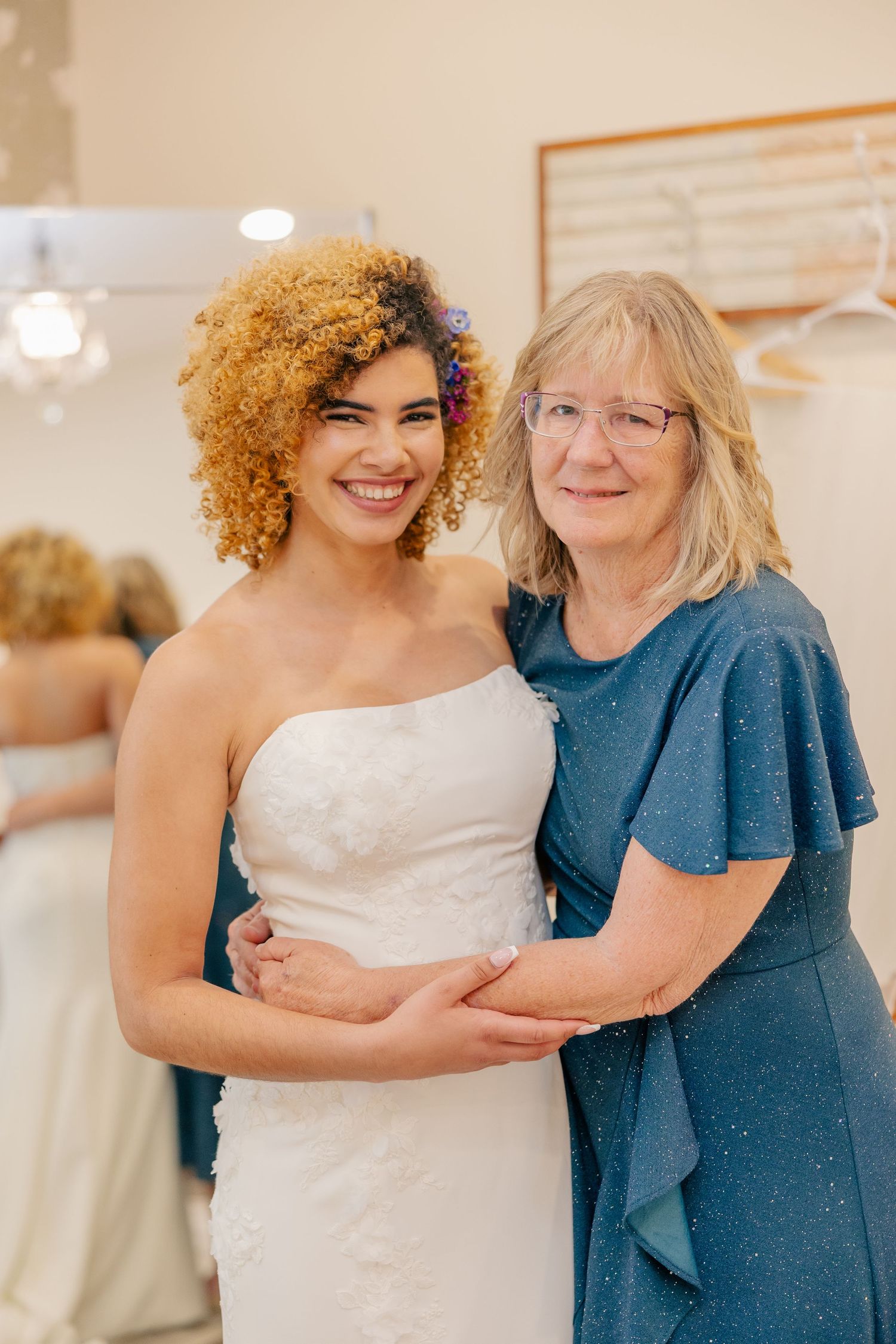 Smiling bride in strapless lace wedding gown poses with older woman in blue dress at bridal shop.