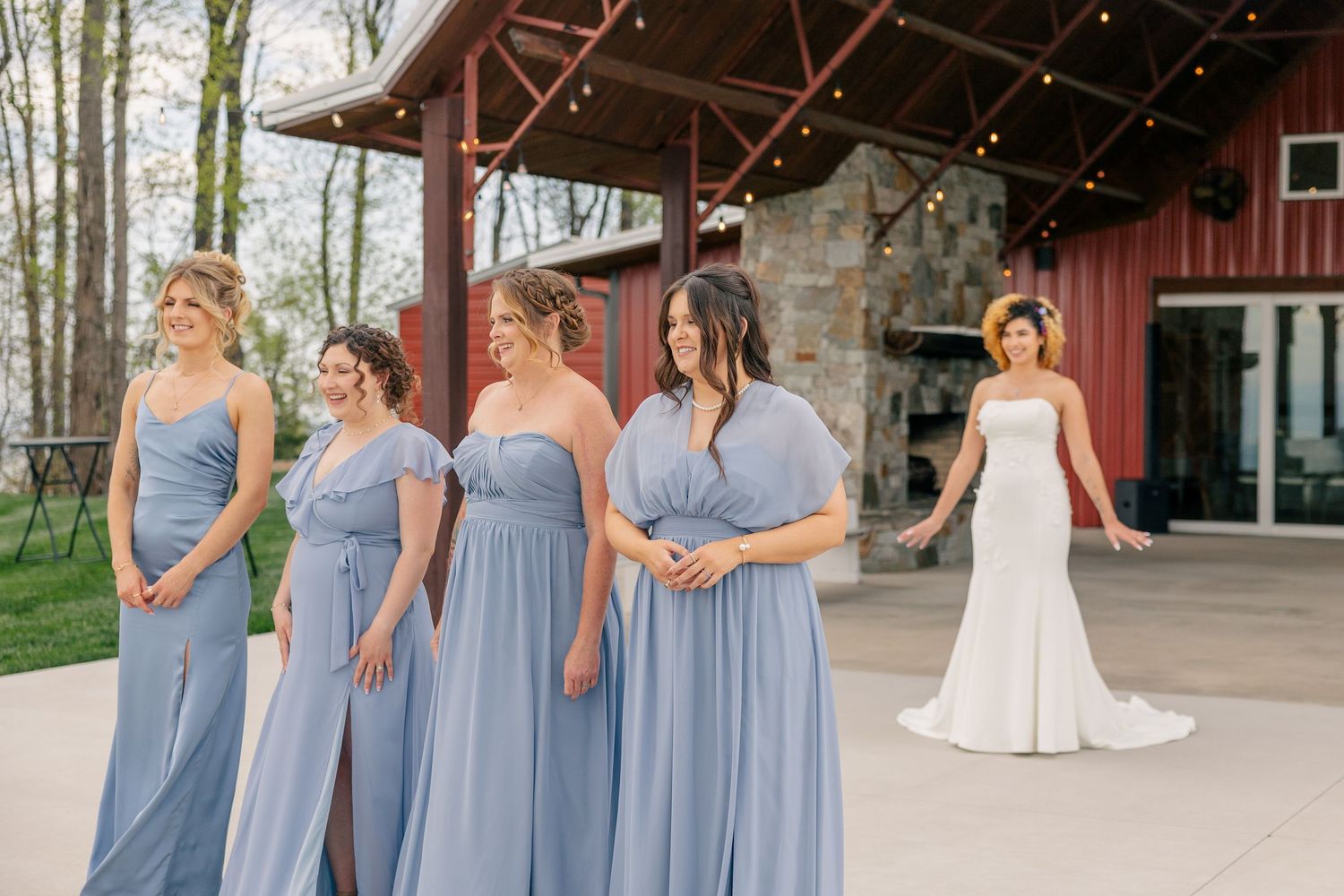 Bride in white gown walks toward bridesmaids in dusty blue dresses outside a rustic barn wedding venue.