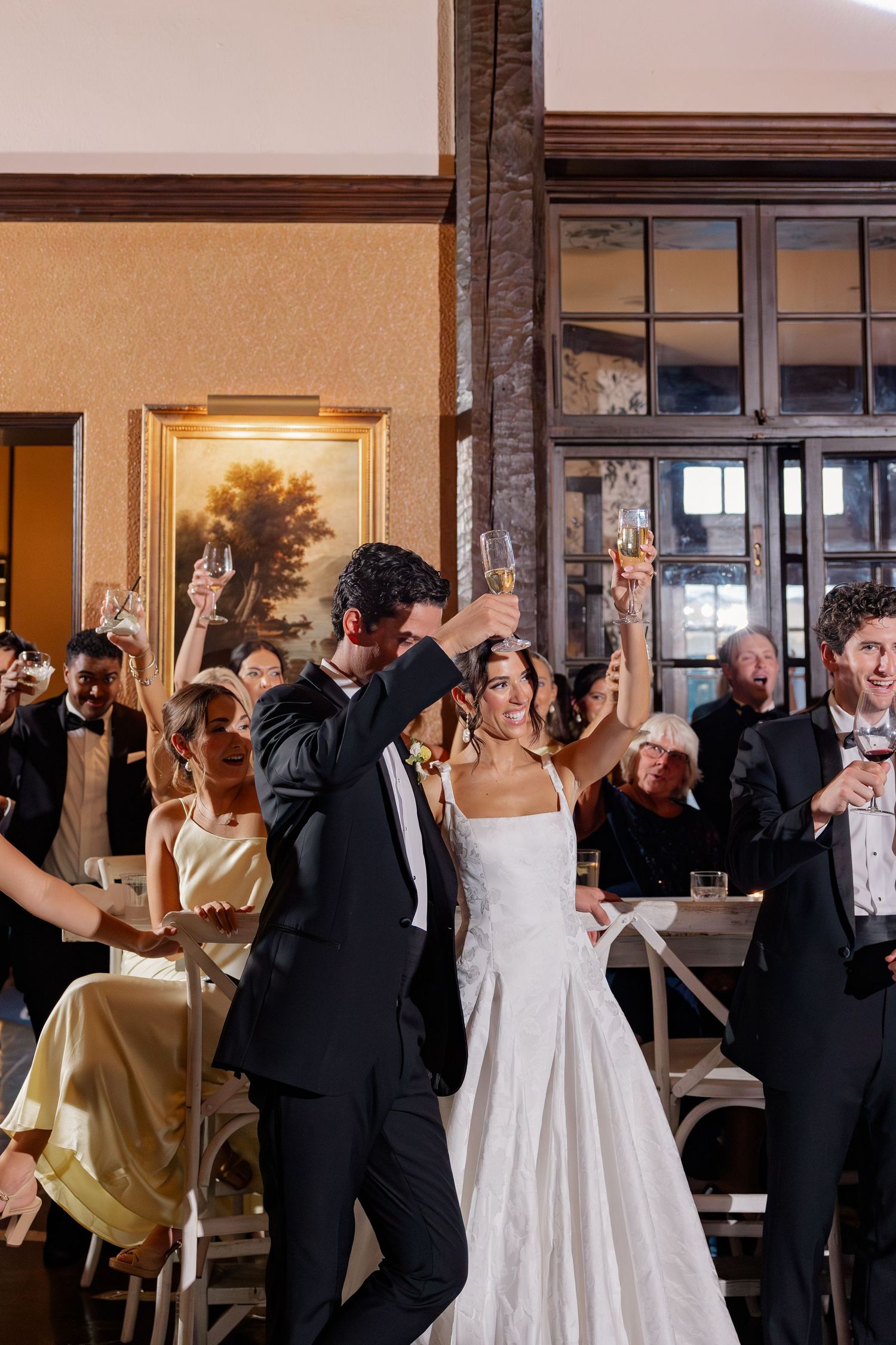 Bride and groom raise champagne glasses with wedding party during celebratory toast at reception.