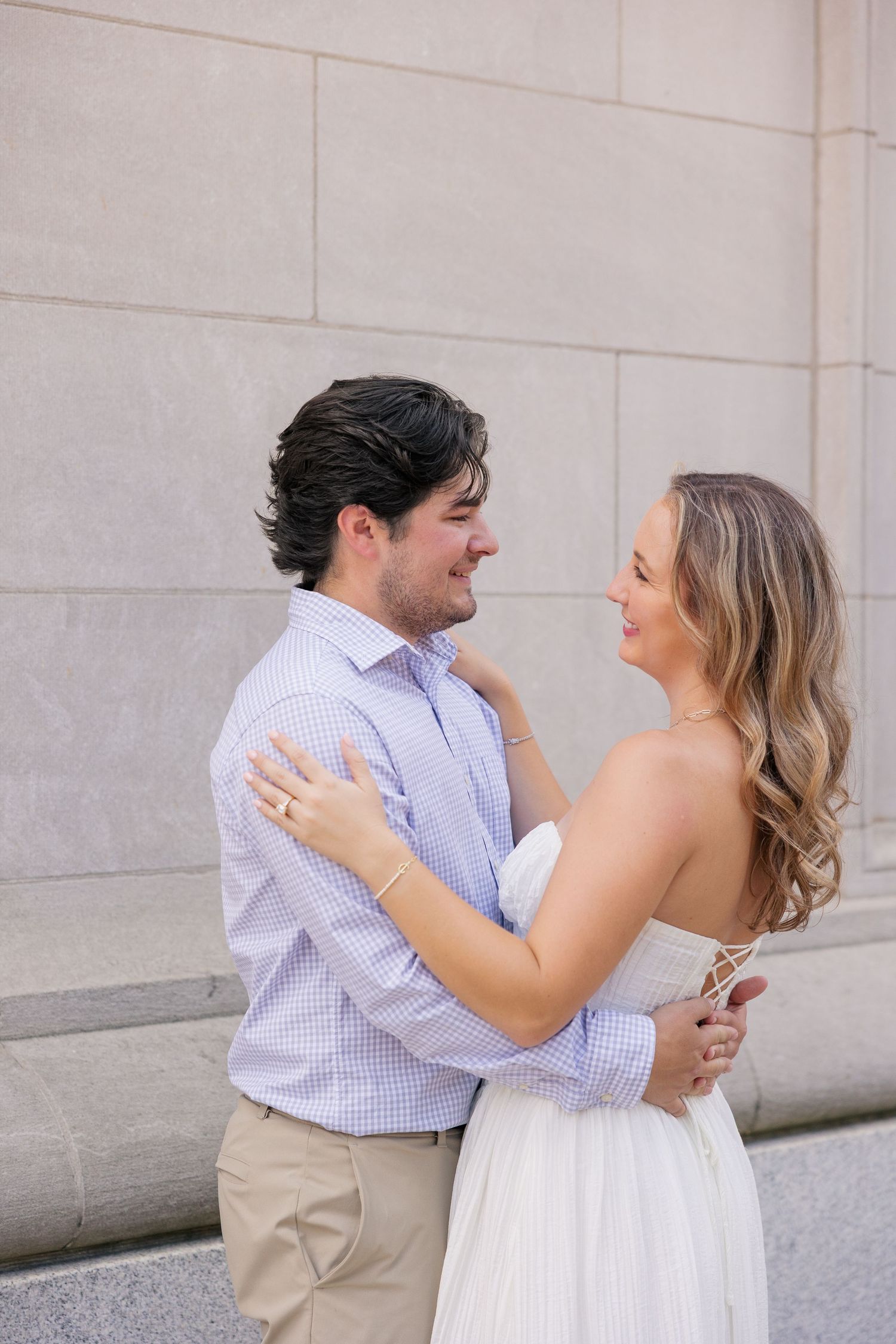Engaged couple embracing and smiling at each other against stone wall, woman in white dress.
