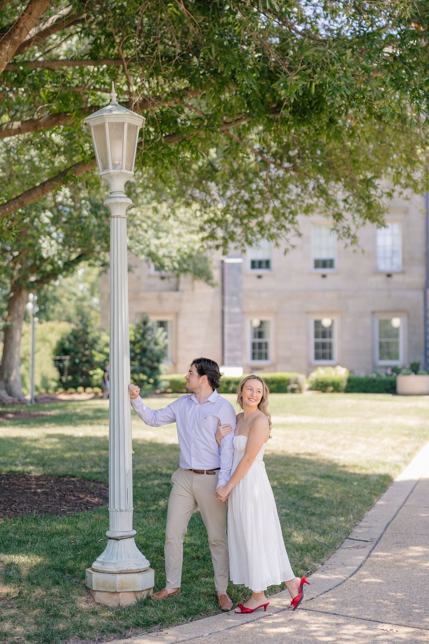 Smiling engaged couple leaning against white lamppost under large tree outside historic building on sunny day.