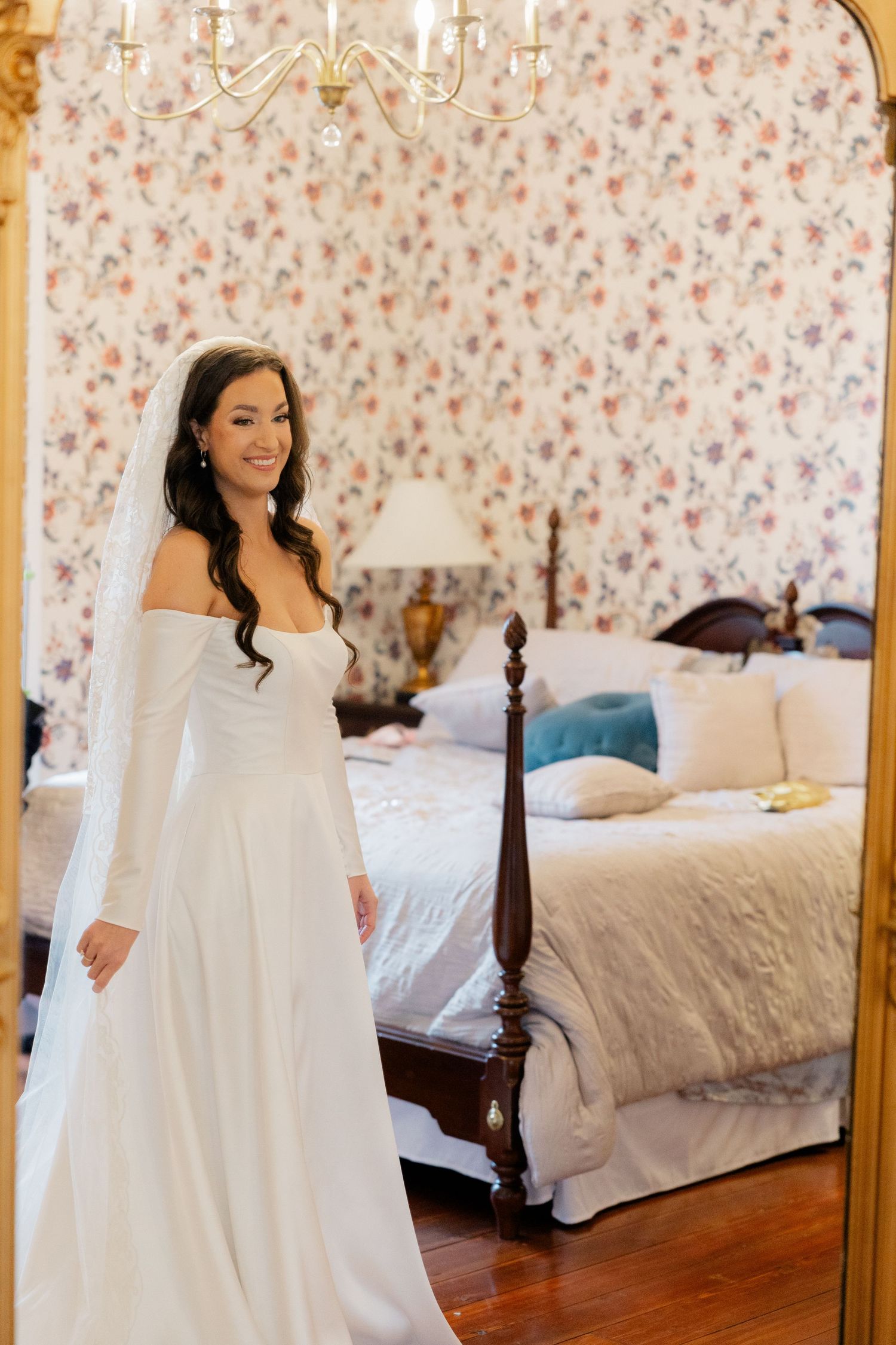 Elegant white wedding gown displayed in a bedroom with floral wallpaper and classic four-poster bed.