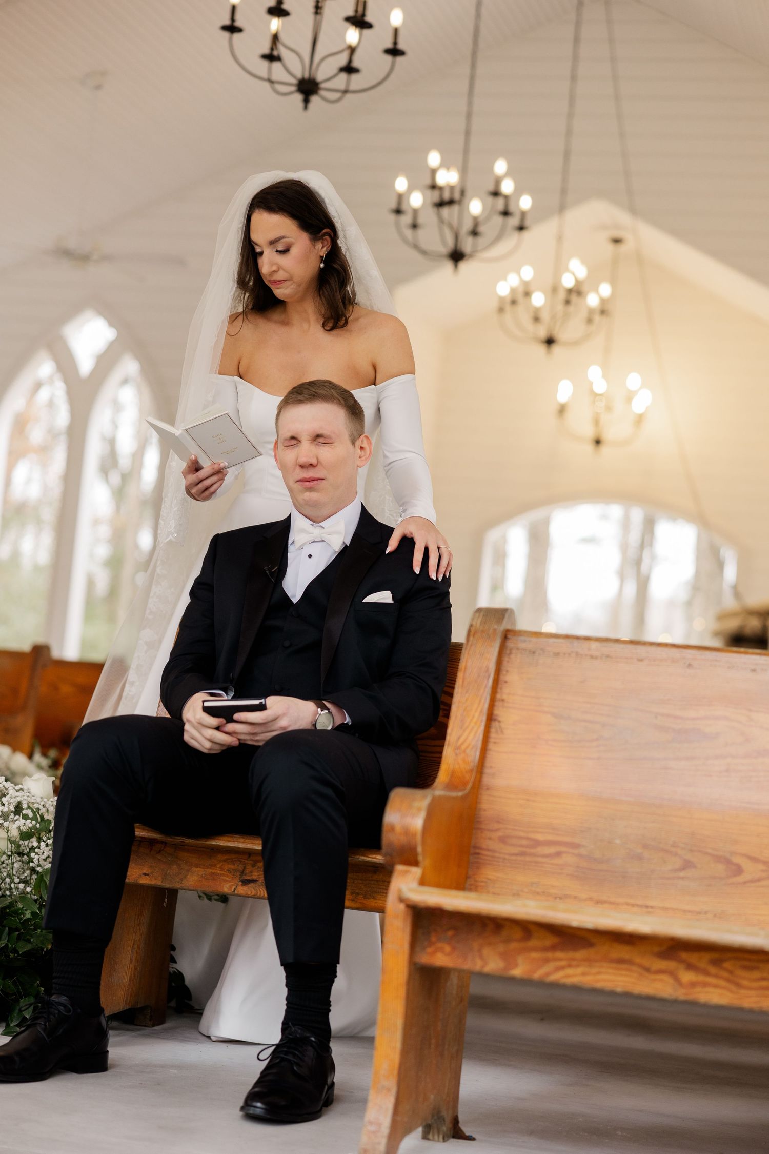 Couple sits together on wooden pews in bright chapel with hanging chandeliers and arched windows.