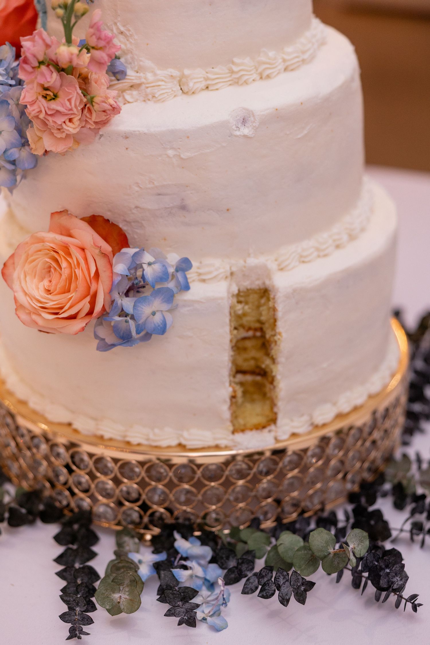 White wedding cake decorated with peach roses and blue delphinium flowers on a textured silver cake stand.