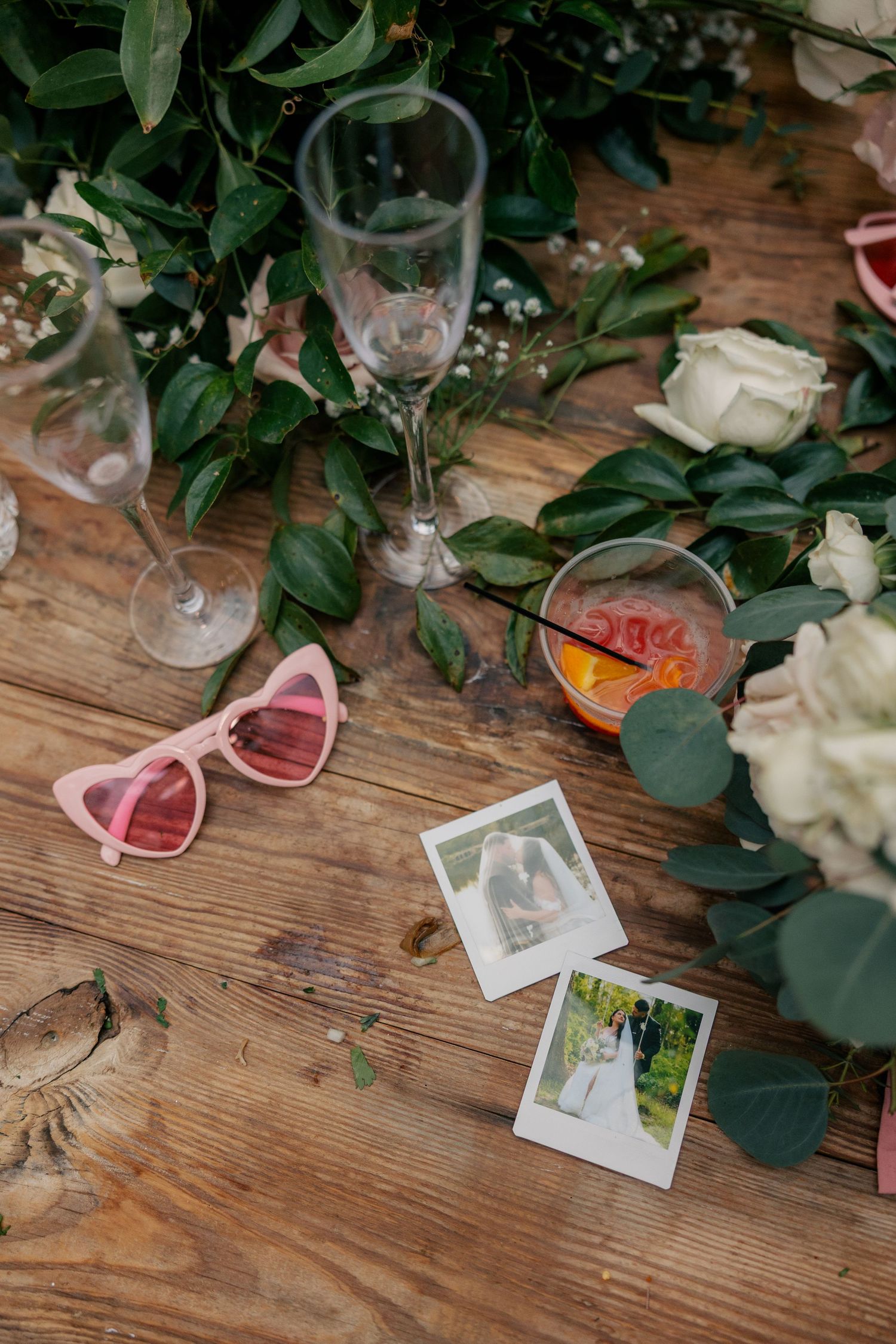 Wedding reception table decor featuring rustic wooden surfaces decorated with green foliage and scattered photographs.