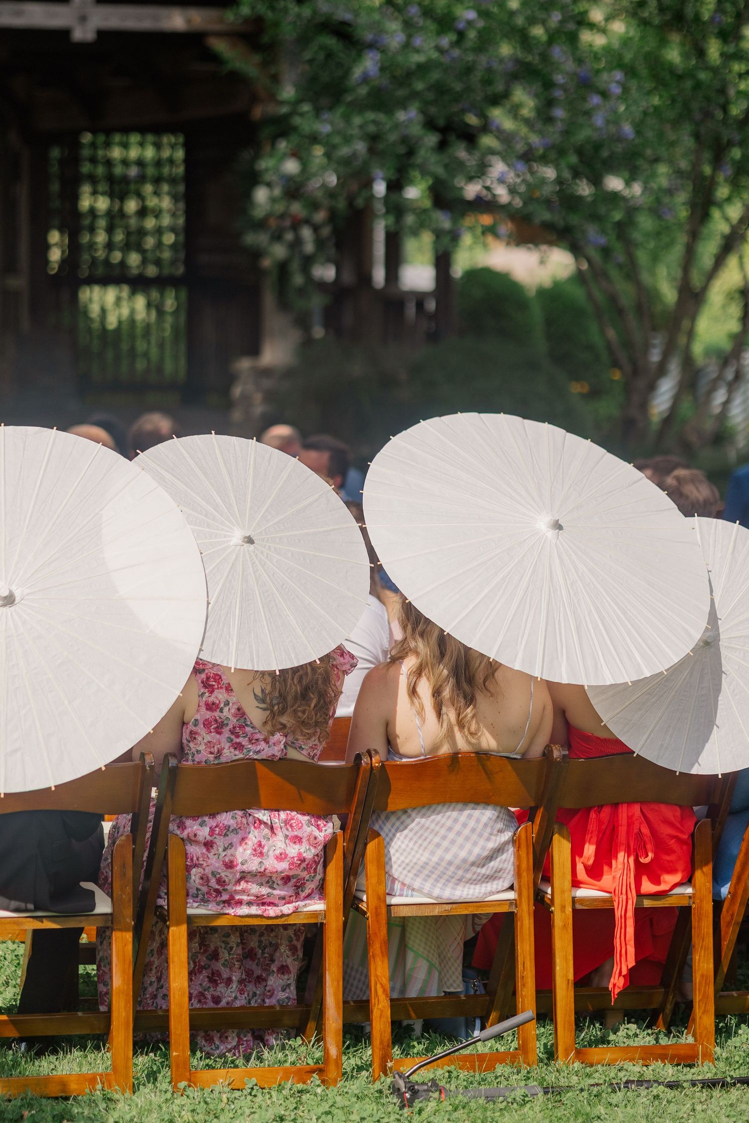 Wedding guests holding white parasols sit in wooden chairs during an outdoor ceremony.