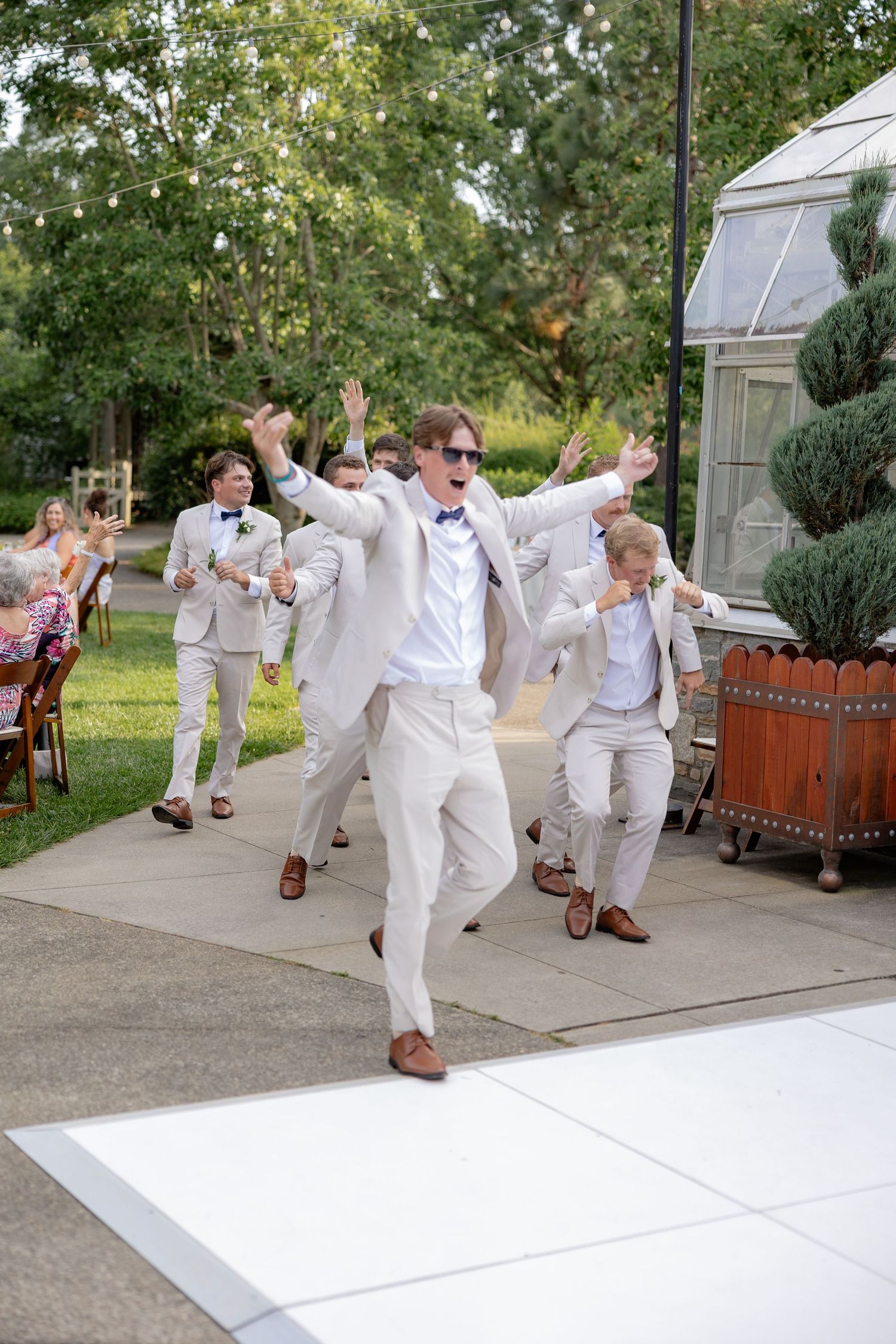 Groomsmen in white suits performing synchronized jumping poses outdoors at a wedding celebration.