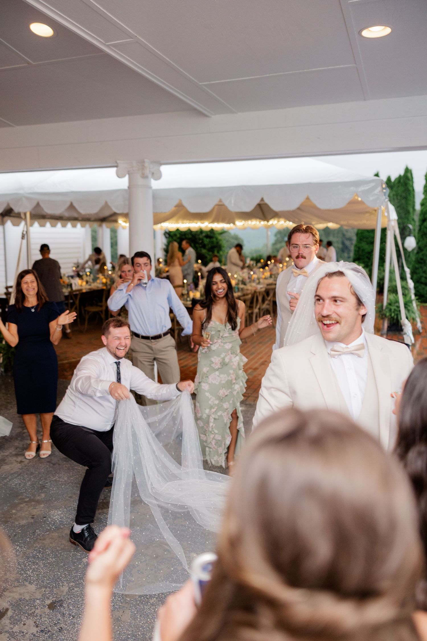 Wedding guests dancing and celebrating energetically under a white tent at an outdoor reception.