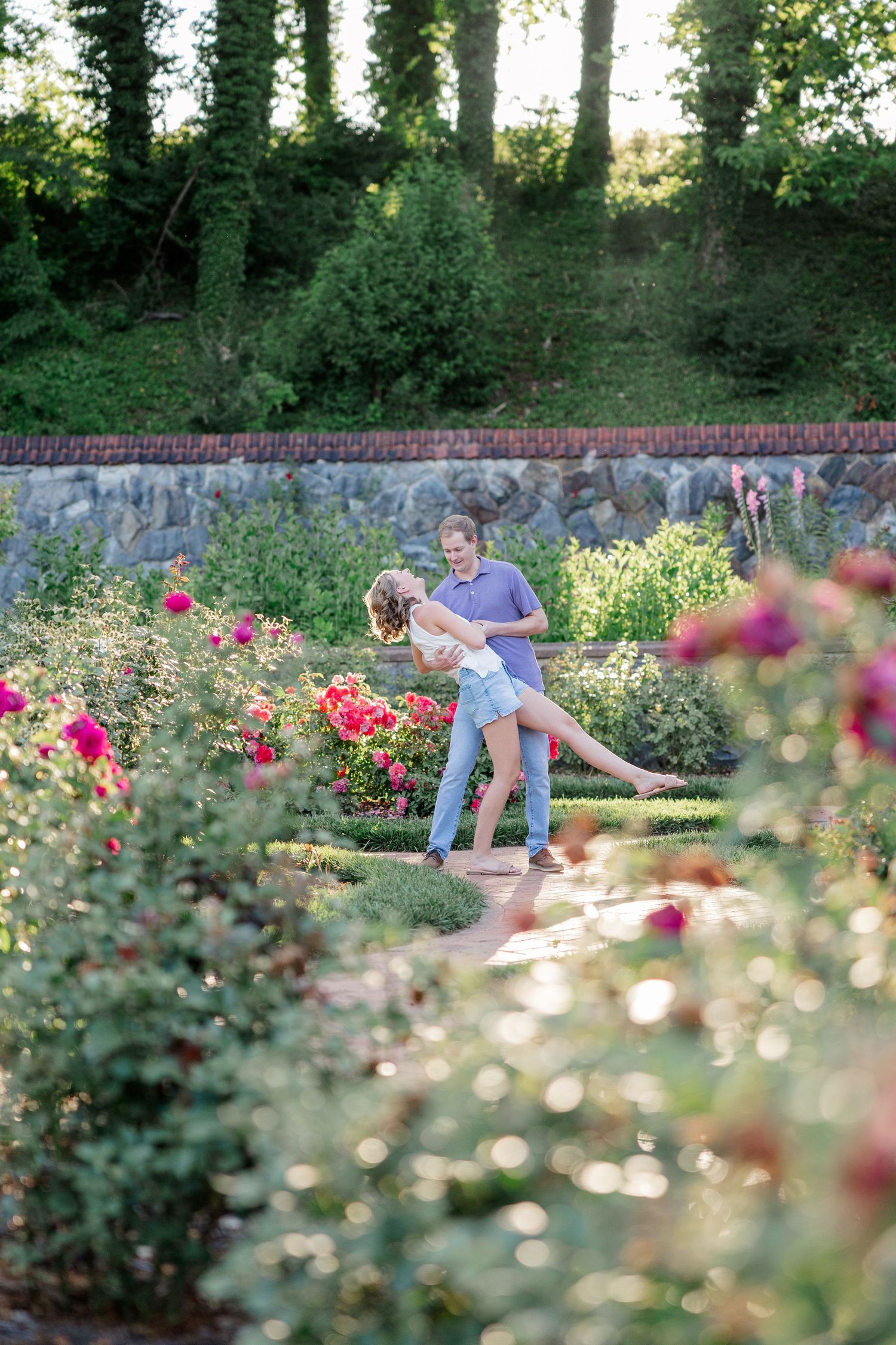 A couple sits casually in a lush rose garden with brick walls and trees in the background.