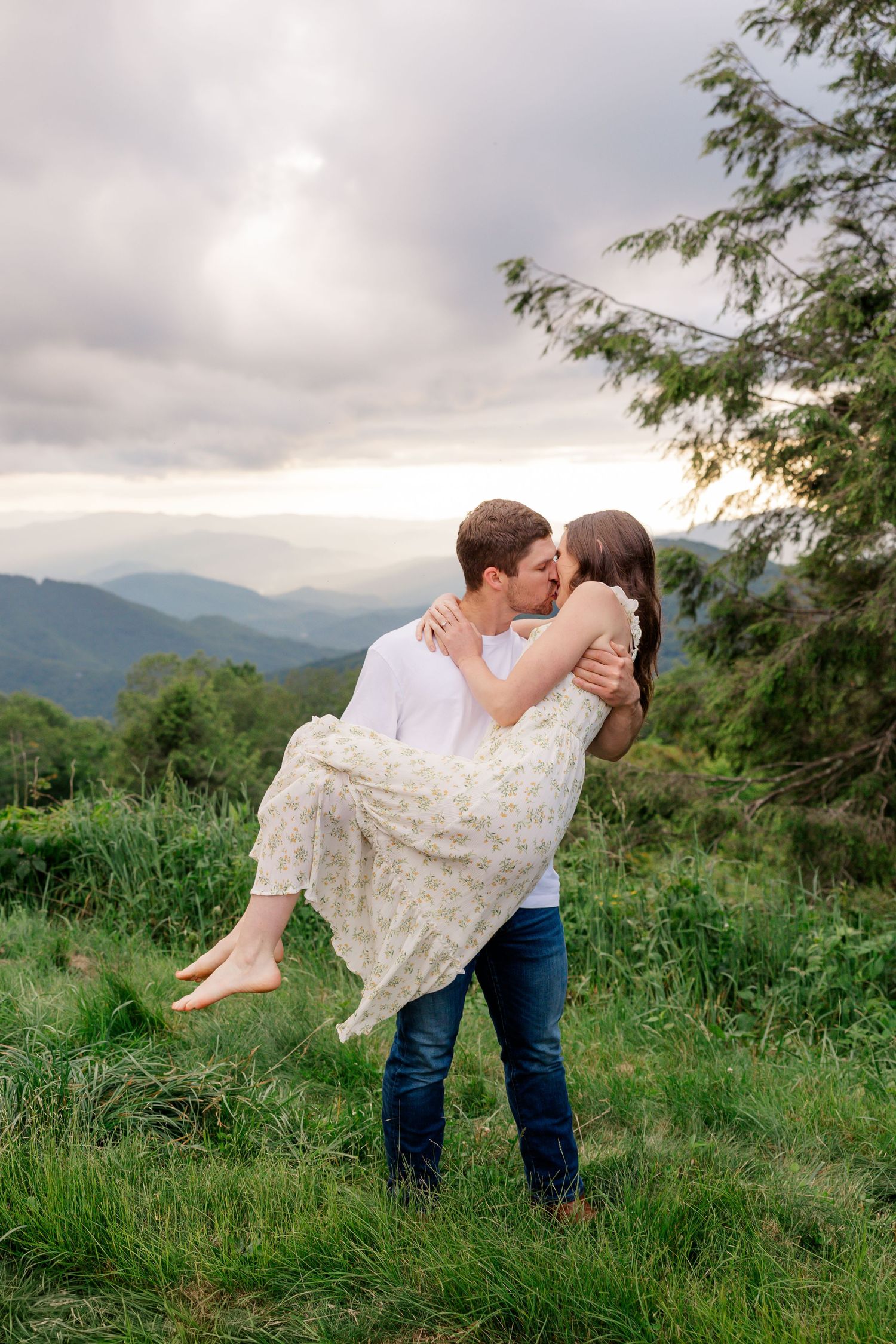 Romantic couple embraces in a mountain meadow with rolling hills and cloudy skies in the background.